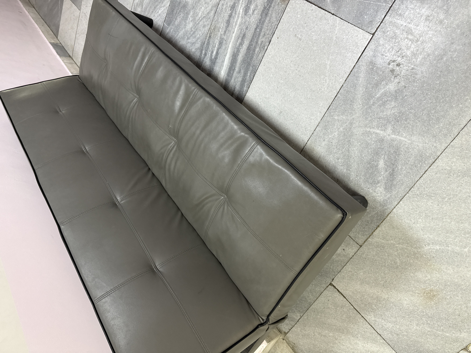 The image shows a gray, tufted, leather couch against a gray tiled wall and floor. A section of a pink surface, possibly a table, is visible on the left. The location is unknown. There are no people visible.