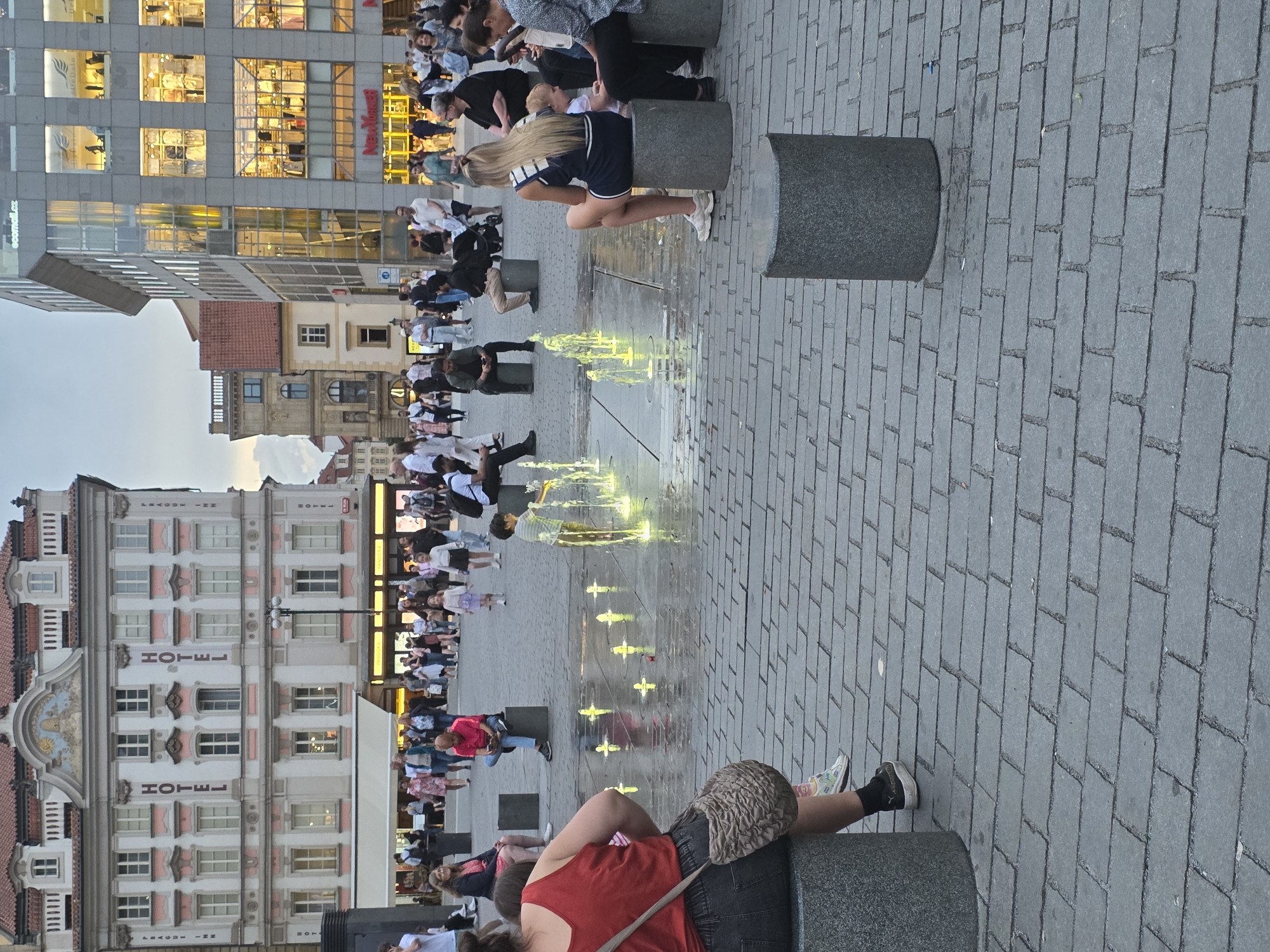The image depicts a lively urban square or plaza during dusk or early evening. The ground is paved with rectangular grey cobblestones, and a linear water fountain with illuminated yellow jets runs through the center, attracting many people.
Numerous individuals, a mix of adults and some children, are gathered in the square. Many are seated on grey cylindrical stone bollards, which serve as informal benches, while others stand or walk around. In the foreground, a woman wearing a red tank top and dark shorts sits on a bollard with her back to the viewer, her hair a light brown, and a woven bag resting on her lap. Further back, a blonde woman in a navy dress with white stripes is also seated on a bollard near the fountain, observing the scene. Other people are scattered throughout the square, some relaxing, others conversing.
The square is flanked by buildings of varying architectural styles. To one side, there's a modern, multi-story building with numerous large glass windows reflecting interior lights. Visible text on this building includes "ecomall.cz" and "Nejlevnejš". On the opposite side stands an older, more ornate building, possibly a hotel, which features the word "HOTEL" written vertically multiple times along its facade, and "LEAGUE INN" also partially visible. The ambient light from the illuminated buildings and the fountain suggests the late evening, though the sky remains a pale grey-blue, indicating the sun has just set or is setting. The overall mood appears calm and social.