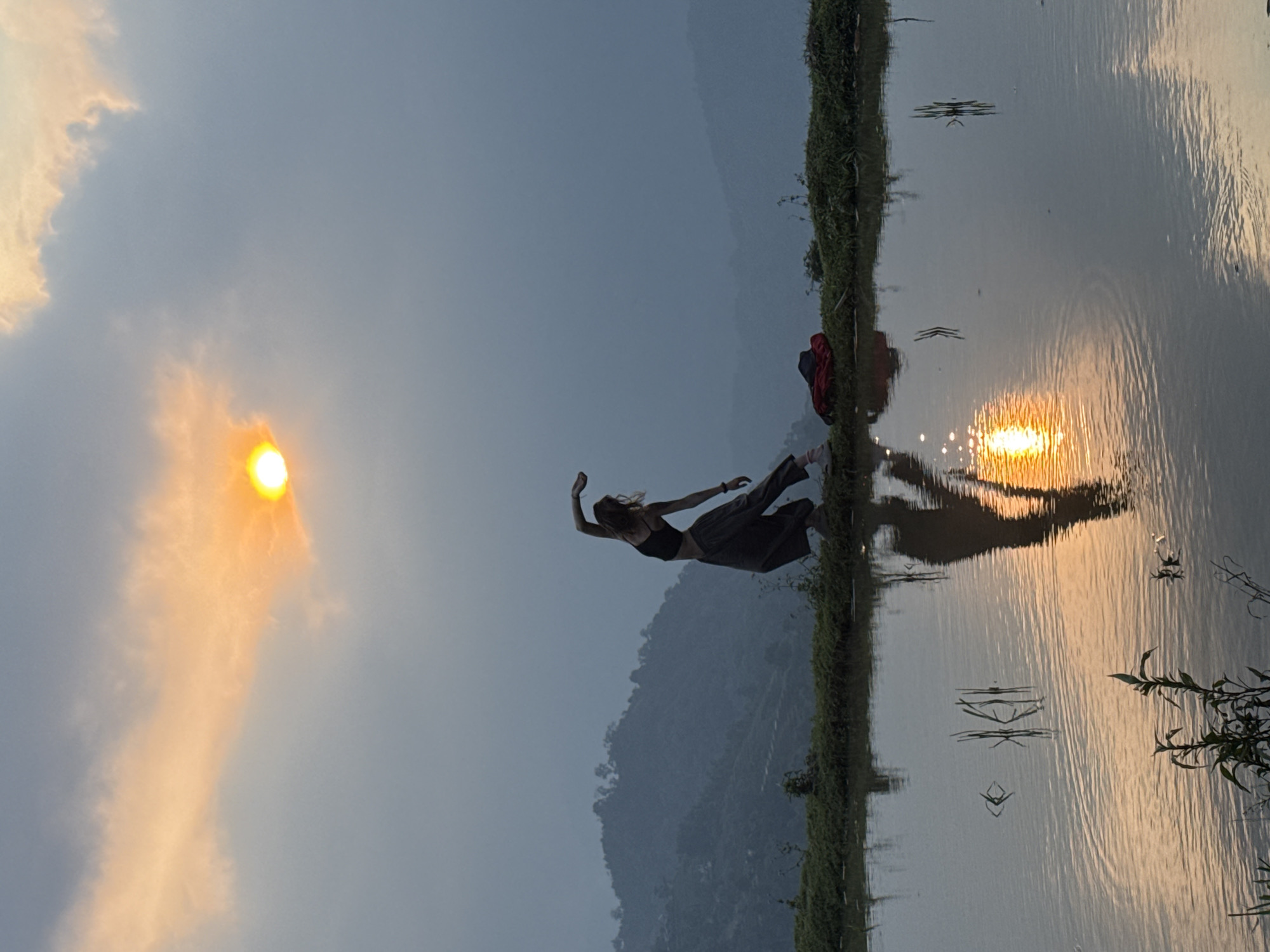 A stunning landscape photograph captured during sunset in Nậm Dịch Commune, Vietnam, depicting a person gracefully suspended in mid-air above a tranquil body of water. The golden hour sun dominates the left side of the frame, bathing the sky in hues of soft blue, orange, and yellow, with light clouds scattered around. Its brilliant reflection creates a shimmering path across the water's surface.
A slender individual, appearing to be female, with long hair, wears a black crop top and dark wide-legged trousers or a skirt. She is captured mid-leap or in a deliberate pose, arms outstretched and legs bent, conveying a sense of weightlessness and joy. This dynamic figure is positioned above a thin strip of lush green grass that borders the placid water, likely a rice paddy field, typical of the Vietnamese landscape. The water beautifully mirrors the sky's colors and the person's silhouette, creating a captivating symmetry. In the reflection, a red bag or backpack is faintly visible on the grassy bank.
Distant, hazy mountains form the serene backdrop, completing the scenic view. The clear weather and soft light indicate dusk. The image evokes feelings of freedom, natural beauty, and peaceful serenity in a rural, mountainous setting. No visible text is present.