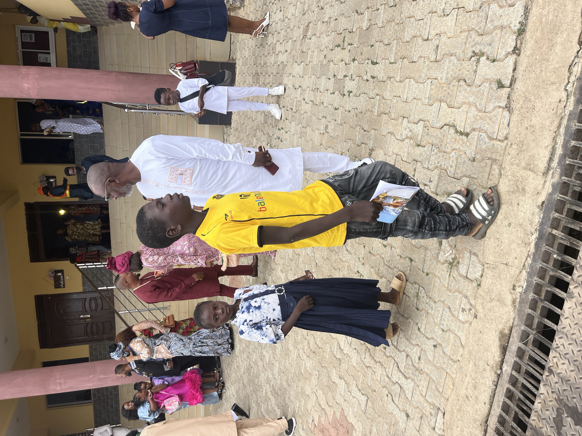 The image depicts a gathering of people outside a building in Abuja, Nigeria, likely a church or event center, given the formal attire and the "Welcome" sign above an entrance. The architectural style of the building, with its colored facade and visible columns, is characteristic of structures found in Abuja. The ground is paved with interlocking concrete blocks.
In the foreground, two children are prominent. A boy in a bright yellow t-shirt and distressed jeans stands facing right, holding a folded booklet. To his left and slightly behind him, a young girl in a blue and white floral top and a dark blue pleated skirt looks directly at the camera. She wears light brown sandals.
Behind them, a group of adults and children are seen, many in traditional African attire. A man in a white kaftan stands to the left of the boy. Further back, people in colorful dresses and suits are interacting. On the right side of the image, a woman in a dark blue dress and white high heels walks away from the camera.
The overall scene suggests an outdoor social event or religious service. The lighting indicates it is daytime. The presence of a metal grate in the lower left corner suggests the paved area is near a drainage system.
