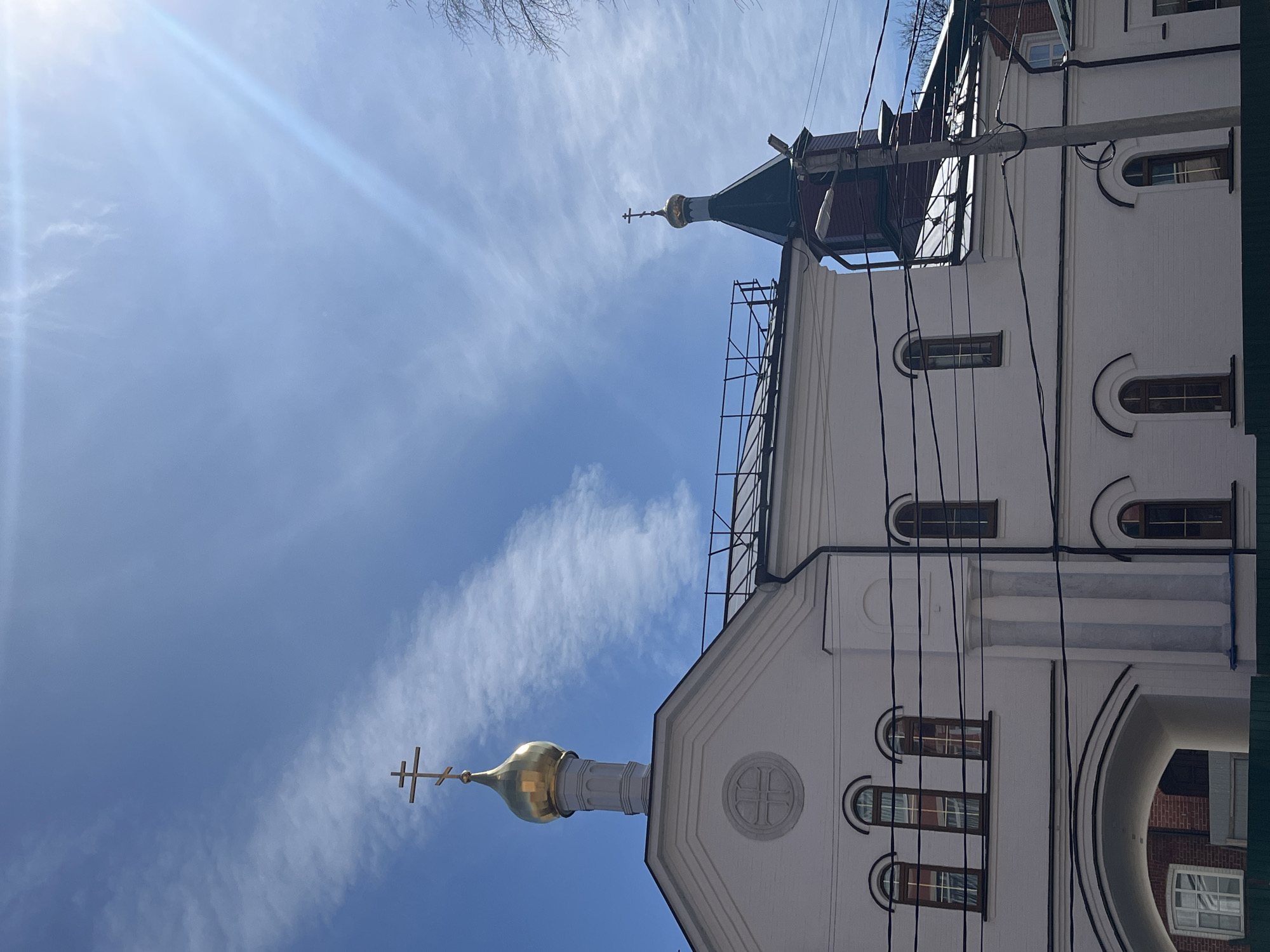 This is an outdoor, daytime photograph of an Eastern Orthodox church or chapel, likely located in Ulyanovsk, Russia, given the context. The image is composed from a low angle, looking upwards towards the building and the sky.
The main subject is a multi-story white building, characterized by its traditional Orthodox architecture. It features two golden onion domes, each crowned with a distinctive three-barred Orthodox cross. The larger dome is prominent on the left side of the frame, while a smaller dome is visible towards the right. The building's facade is painted white, with decorative architectural elements and several arched windows, some of which are partially obscured.
The sky is a clear, bright blue with scattered, wispy white clouds. A strong sun glare emanates from the upper left corner of the frame, indicating bright sunshine. Bare tree branches are visible at the very top edge of the image. Numerous dark utility cables or power lines run across the scene, particularly dense on the right side, adding an urban element to the setting. Scaffolding can be seen near the roofline and the smaller dome on the right, suggesting ongoing construction or maintenance. A dark archway at the bottom right reveals a glimpse of a red-brick building behind the main structure.
No people are visible, and there is no discernible text within the image. The time of day is clearly daytime, characterized by bright sunlight and clear weather.