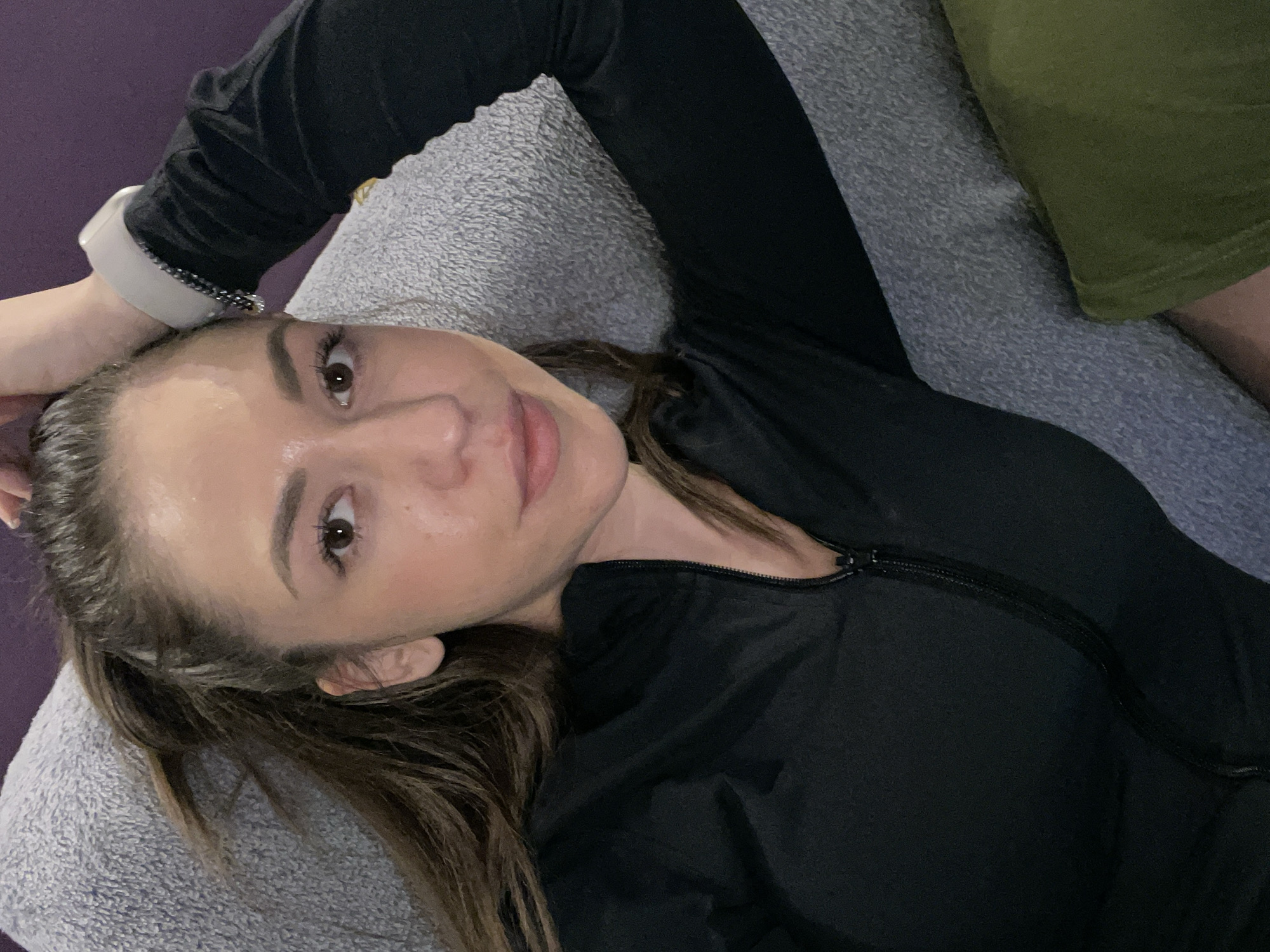 A woman is lying down on a grey, textured surface, possibly a couch or cushion. She is wearing a black, long-sleeved zippered top. Her hair is brown and pulled back, and she is wearing a white smartwatch on her wrist. Part of a green garment is visible in the upper right corner of the frame. The background is a muted purple. The location cannot be determined as Sofia, Bulgaria based on visual evidence.