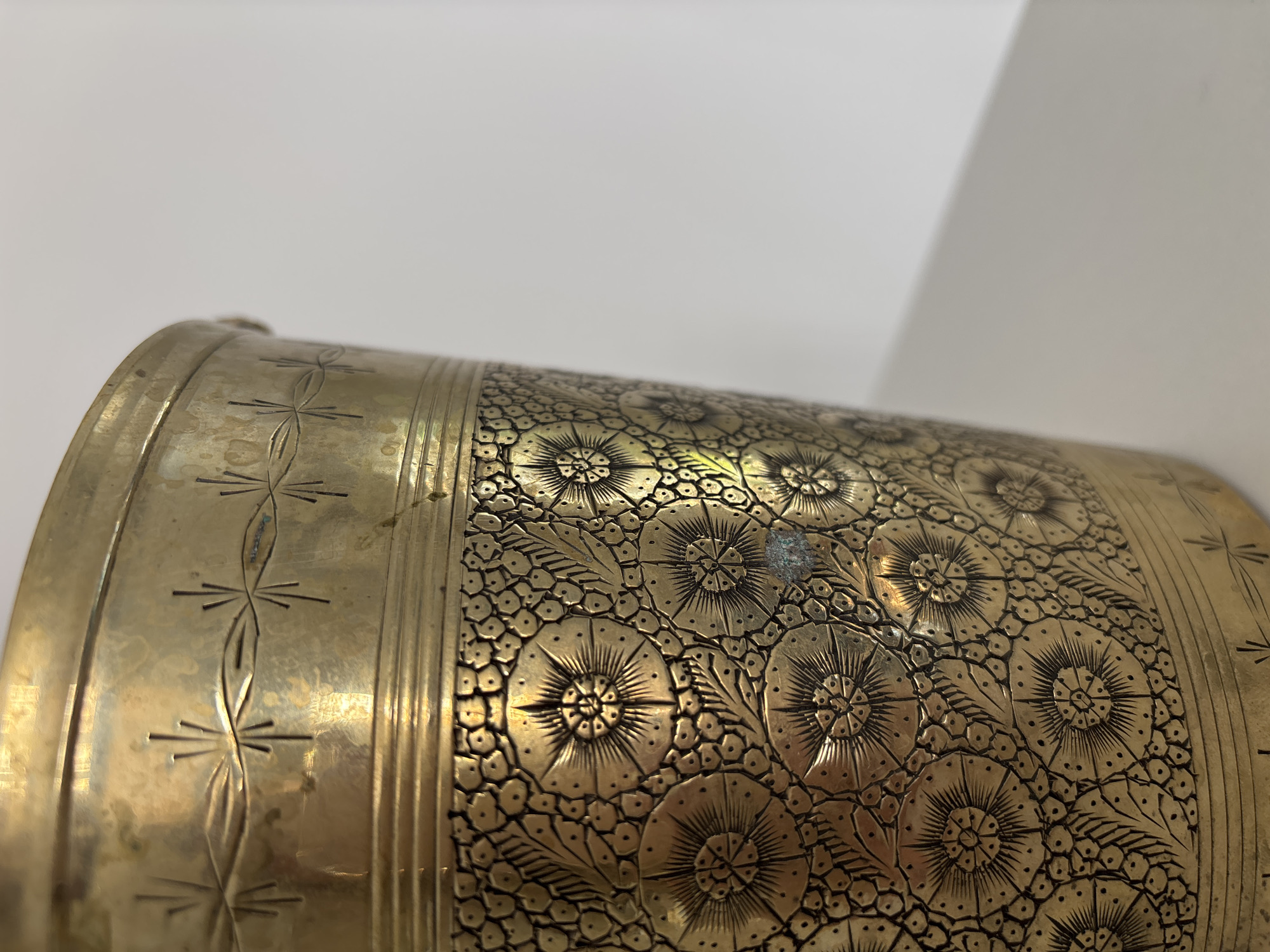 The image shows a close-up of a brass cylindrical vessel with intricate floral and geometric etchings. The vessel is positioned in a studio setting, with a plain white background and a light-colored surface beneath. The brass has a warm, golden sheen and exhibits some signs of age and wear, including minor discoloration. The etchings are detailed, featuring repeating patterns of flowers with radiating petals and stylized leaves, bordered by bands of linear designs. The lighting highlights the embossed textures of the metal. The location in Razdolnoye, Russia, is not discernible from the visual content of the image.