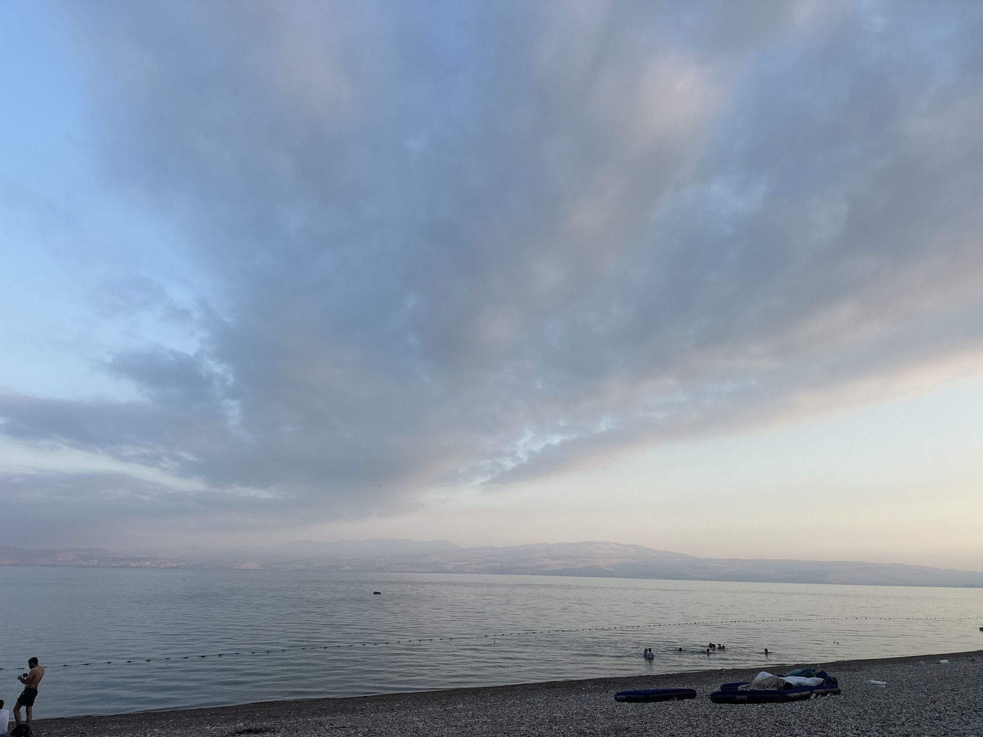 This image depicts a view of the Sea of Galilee in the Emek HaYarden Regional Council, Israel. The scene is a pebble beach with calm water extending towards a distant shoreline. The sky is filled with scattered clouds, predominantly light blue and gray, with a soft glow indicating either early morning or late afternoon. In the foreground, a few individuals are visible on the beach and in the water. One man stands on the left side of the beach, looking towards the water. Further into the water, several people are swimming or wading. On the beach, there are inflatable mattresses and some personal belongings. The distant landscape across the sea consists of low-lying hills and mountains shrouded in a light haze. A rope barrier is visible in the water, marking a swimming area. The overall atmosphere is serene and quiet.