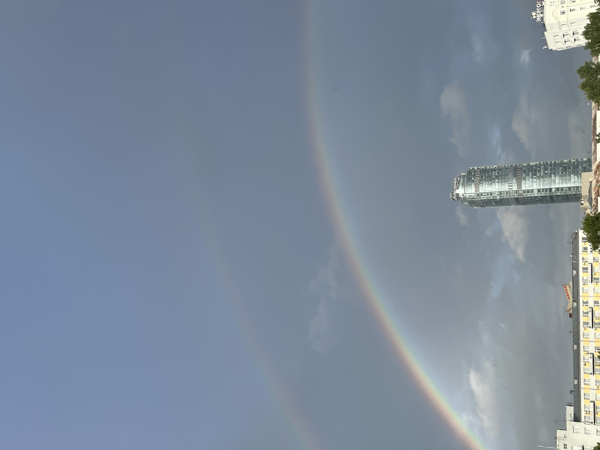 The image captures a cityscape under an overcast sky featuring a double rainbow. A prominent primary rainbow arcs from the lower-left to the upper-right, with a fainter secondary rainbow visible parallel above it, particularly on the left. The sky is largely covered with dark grey clouds, especially on the right and in the lower half of the frame.
In the mid-ground, a tall, modern skyscraper, identified as the Iset Tower, stands prominently on the right side. Its structure is cylindrical or slightly tapered, clad in reflective glass panels, and features a distinctive, multi-tiered top.
In the lower-left foreground, a multi-story building with a white facade and numerous rectangular windows is partially visible. Adjacent to it, a segment of a yellow building with white-framed windows is seen, featuring multiple external air conditioning units and a red sign with the Cyrillic text "АНТЕН" (ANTEN). Further to the right, partially visible on the far right edge, is a white building with "ЭНЕРГОГ" (ENERGOg) in large Cyrillic letters on its roof, which is consistent with the House of Soviets or a nearby building.
Green tree foliage is visible at the very bottom of the frame, obscuring the lower parts of the foreground buildings. No people or specific actions are depicted in the scene.
The location is identifiable as Yekaterinburg, Russia, with the Iset Tower (Башня Исеть) and the surrounding architecture confirming the urban environment. The perspective is likely from a southwest or west vantage point, looking towards the northeast or east, capturing the rainbows against the stormy sky above the city landmarks.
