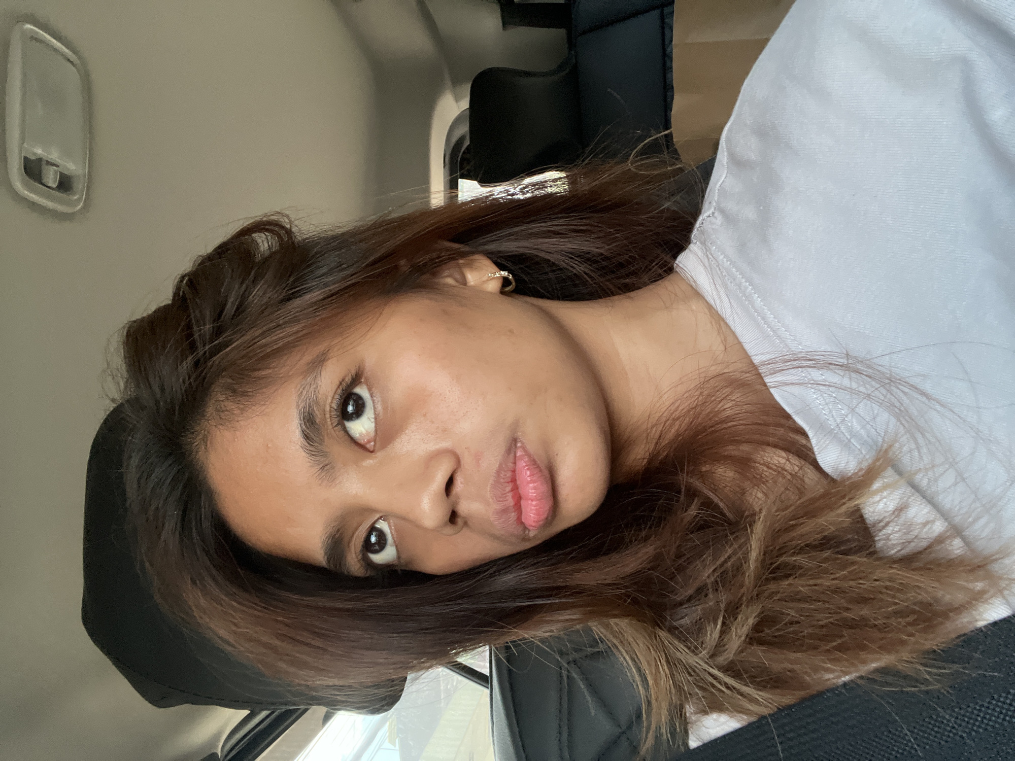 A young adult female is depicted seated inside a vehicle, likely the front passenger seat, in Davao City, Philippines. She has long, dark brown hair with visible lighter highlights or balayage, parted on her left, falling over her shoulders. Her complexion is medium-dark, and her brown eyes are directed upwards. Her lips are distinctly pursed into a pout. She wears a plain white short-sleeved t-shirt and small gold hoop earrings adorned with minute stones. The car's interior features black, possibly leather, seats with a quilted pattern on the visible headrest. The ceiling is light grey with an overhead light fixture. A brown paper bag is present on the seat to her right. The exterior through the window is blurred, showing hints of green foliage and structures, indicative of an urban or suburban environment.