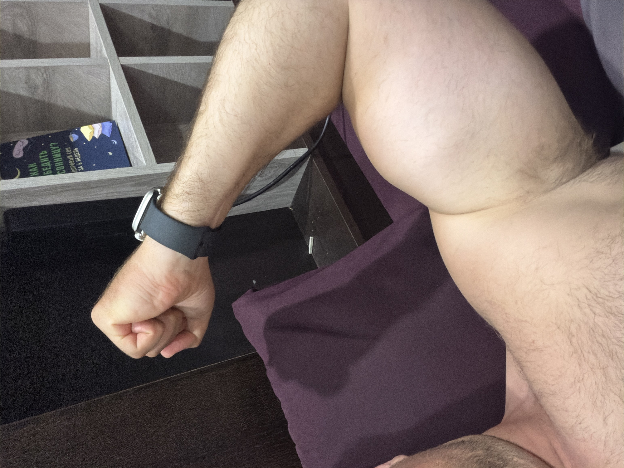A close-up shot captures a man's bicep flexed, with his forearm and wrist visible. He is wearing a dark smartwatch on his left wrist. The background features a bookshelf with a book titled "КАК БЕДИТЬ СОЛНИЦУ?" and a purple piece of fabric, possibly upholstery. The scene appears to be indoors, with a dark wooden surface and a light-colored wall in the background. No specific location in Baan Mae Nam, Thailand, can be determined from the image.
