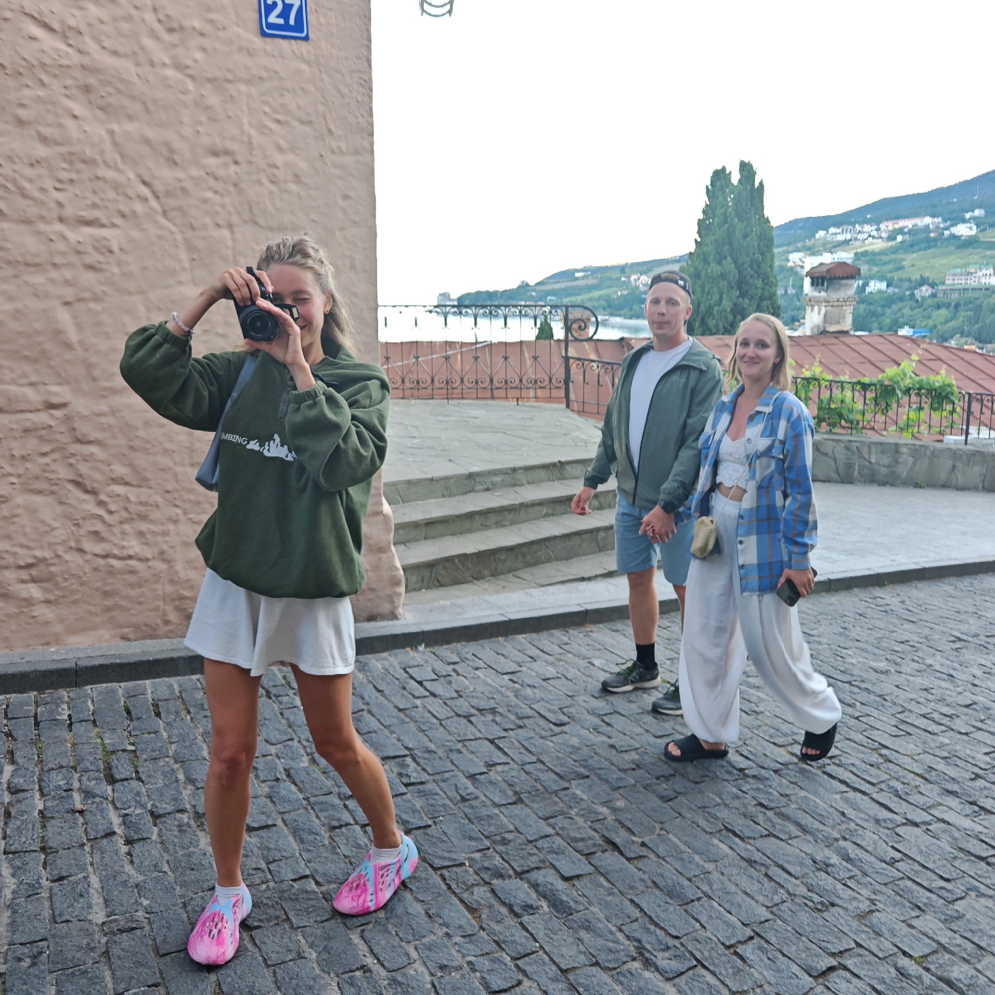 Three people are walking on a cobblestone street. The person in the foreground on the left is a young woman with blonde hair, wearing a green hoodie and white shorts. She is holding a camera up to her face, appearing to take a picture. She is wearing bright pink and blue patterned shoes. To her right, a couple walks hand-in-hand. The man is wearing a green jacket over a white t-shirt and blue shorts. The woman is wearing a blue and white checkered shirt over a white top and loose white pants. She has a small beige purse slung over her shoulder and is holding a black phone in her right hand.
The background shows a glimpse of a town on a hillside overlooking a body of water, possibly the sea. Buildings with red tile roofs are visible, nestled amongst green foliage and trees. There are stairs leading up from the street behind the woman with the camera. A sign with the number "27" is visible on a light-colored building wall on the left. The scene appears to be outdoors during the daytime, with overcast skies. The mood seems casual and relaxed, suggesting a tourist outing.