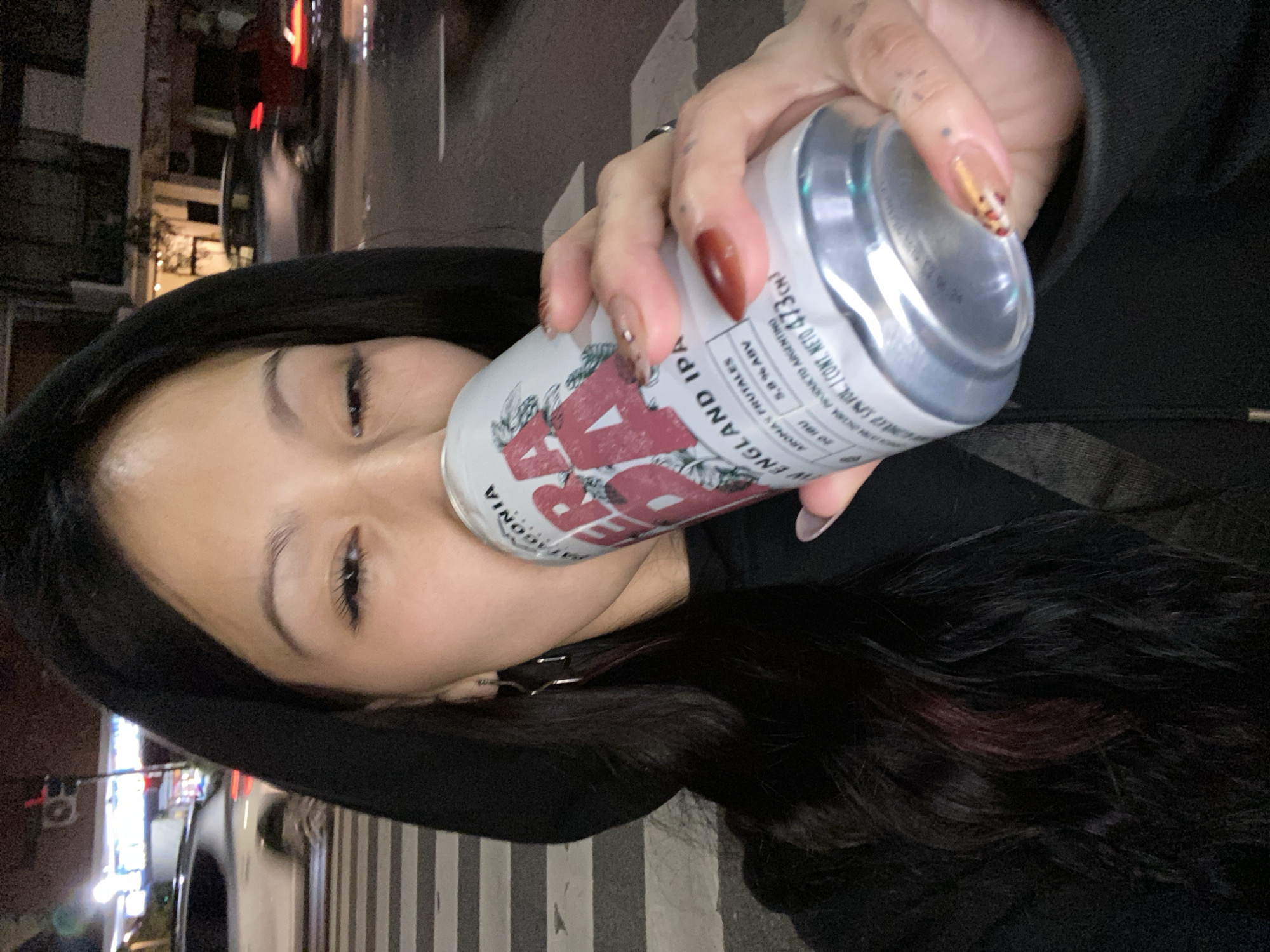 A woman in a black hooded sweatshirt is drinking from a white aluminum can labeled "Patagonia England IPA, aromas frutales, 5.8% ABV, infol Cont. Neto 473 cm." The can's label also prominently displays "Vagonia RA" with a triangular symbol. She holds the can with her right hand, which has manicured nails with varying colors and designs. Her left side of her face and part of her long, dark hair (with a hint of burgundy) are visible. The location is outdoors, seemingly at night, next to a city street in Buenos Aires, Argentina, where traffic and buildings are visible in the background. The sidewalk has striped markings.