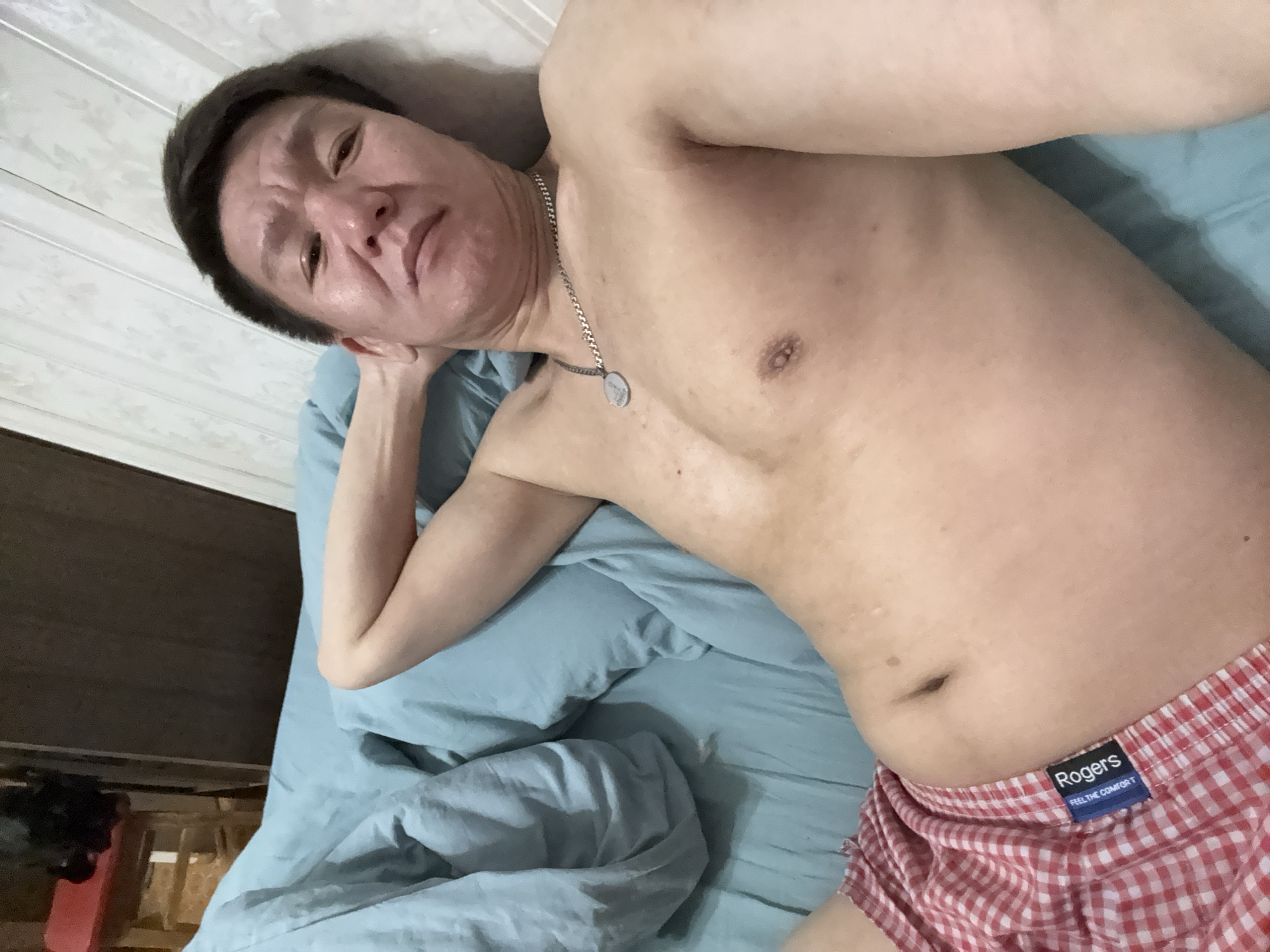A shirtless Asian man wearing red and white checkered boxer shorts with the brand name "Rogers" visible on the waistband is lying on a light blue bedsheet. He is wearing a silver chain necklace with a pendant. The man is facing the camera with a neutral expression. His head is resting on his left hand. The background shows a partially visible wall and furniture, suggesting an indoor setting. The location is not discernible based on visual information.