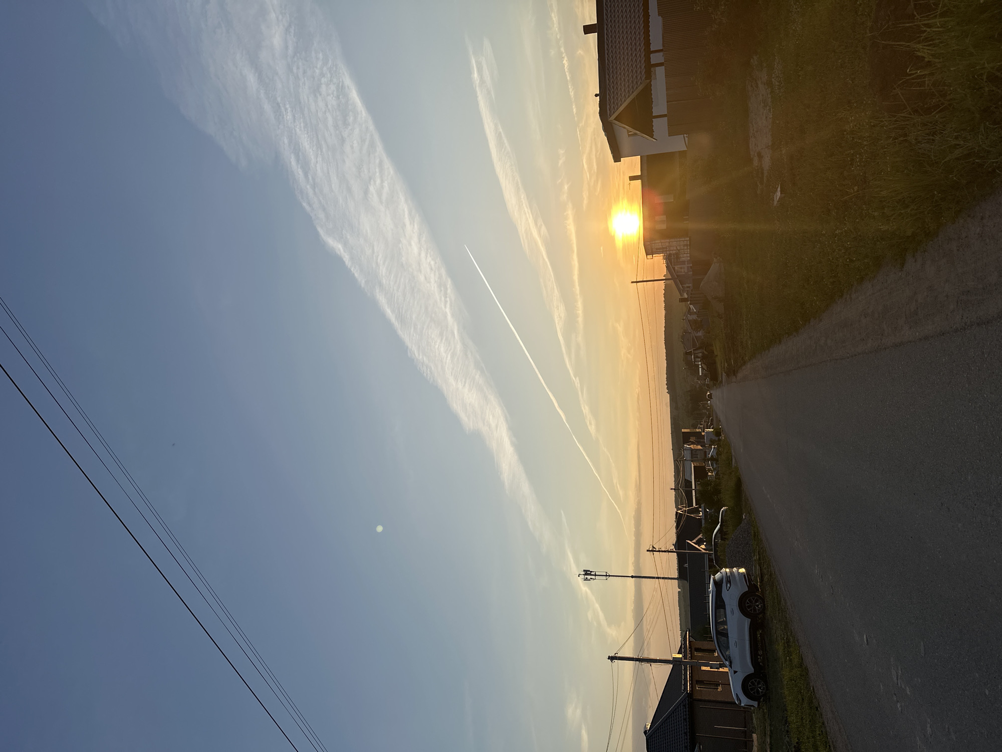The image depicts a rural road in the village of Estachi, Russia, during sunset. The sun, a bright orb, is low on the horizon, casting a warm glow and long shadows. The sky is a gradient of blue and orange, with streaking clouds, possibly contrails from an airplane. Several houses with dark roofs line the road, some appearing to be under construction. A white SUV is parked on the left side of the road, near a house. Power lines stretch across the sky, supported by utility poles, one of which is a cell tower. Vegetation, including grass and small trees, grows along the roadside and around the houses. The overall scene is peaceful and evokes a sense of a quiet evening in a semi-rural setting.