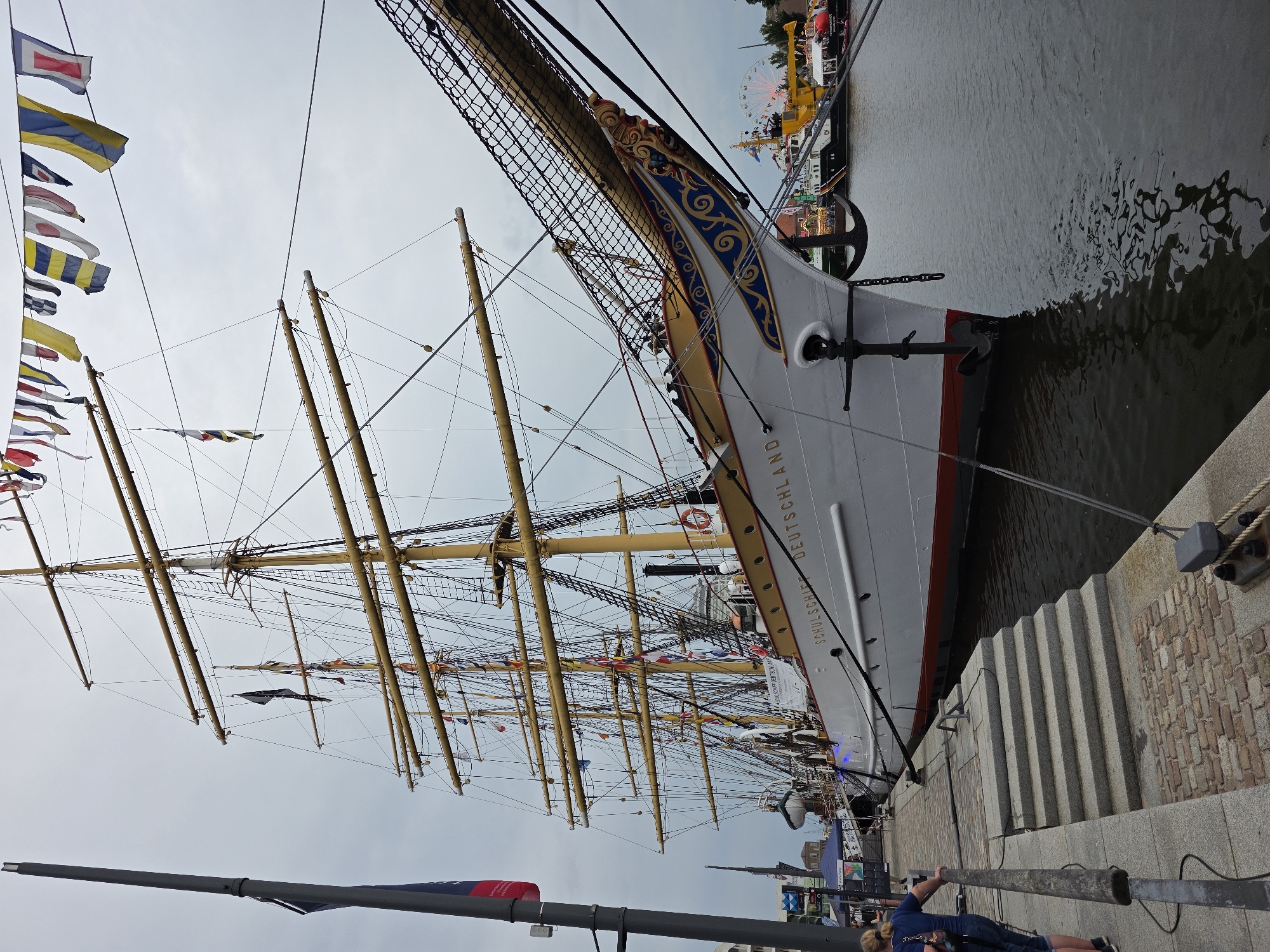 A large, white three-masted sailing ship named "SCHULSCHIFF DEUTSCHLAND" is docked at a waterfront in Bremerhaven, Germany. The ship is adorned with numerous flags, including a string of international signal flags strung between its masts and a German flag on a pole at the bow. An anchor hangs from the bow, partially submerged. The ship is tied to a stone quay where a person wearing a blue shirt and jeans is standing, leaning on a railing. Behind the ship, other boats and a Ferris wheel are visible in the harbor. The sky is overcast.