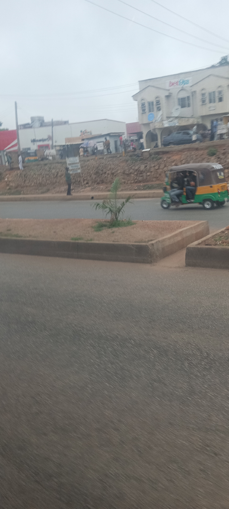 This street scene in Jos, Nigeria, depicts an urban environment under an overcast sky. In the foreground, a grey asphalt road shows signs of motion blur, suggesting the image was captured from a moving vehicle. A concrete median strip runs along the road, featuring a small, green plant and some sparse grass.
In the mid-ground, a green and yellow motorized tricycle, commonly known as a Keke Napep, is seen driving on the road with several passengers inside. To the left of the median, a person stands beside a low, rocky embankment that separates the road from the buildings in the background.
The background features a row of commercial buildings. A prominent two-story white building stands out, displaying the red and white "bet9ja" logo on its facade. Other structures, including one with a red roof, are partially visible. A dark-colored car is also seen near the buildings. Several people are scattered throughout the background, engaged in various activities like walking or standing. Overhead, utility poles and power lines are visible against the bright, hazy sky. The scene captures typical daily life on a street in a Nigerian city.
