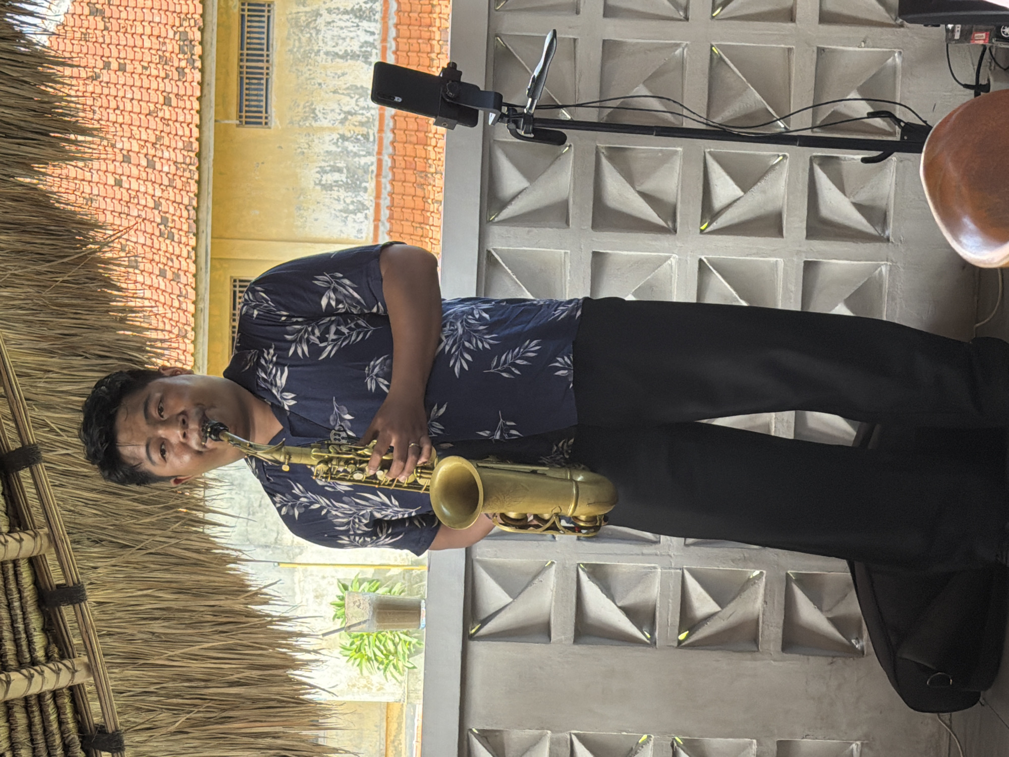The image captures a man playing a saxophone in Kuta Selatan, Indonesia. He is wearing a dark blue shirt with a leaf pattern and black pants. He is positioned in front of a wall with square, geometric cutouts. To his left is a thatched roof structure. In the background, there is a building with a red-tiled roof. A mobile phone attached to a stand is positioned above him. A brown drum is visible near the top right corner.