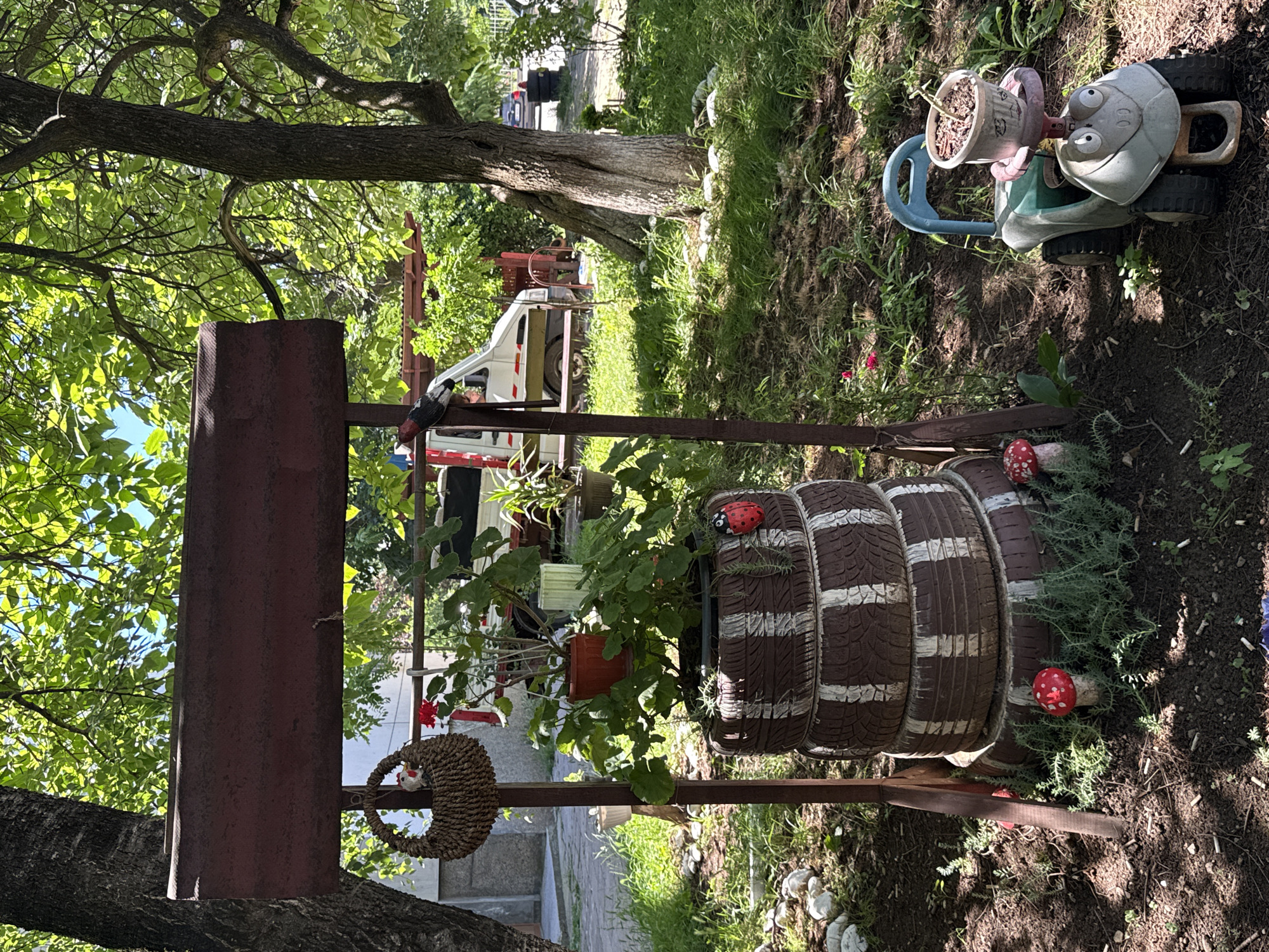 The image depicts an outdoor scene, likely a garden or yard, in Vratsa, Bulgaria. In the foreground, a rustic structure resembling a well-head is constructed from metal and wood. A basket hangs from one of the posts. Below this structure, a tiered planter is made from stacked tires, painted with brown and white stripes, holding green plants. Decorative elements include a ladybug figurine on the tires and two red and white mushroom figurines at the base. To the right of the planter, a child's ride-on toy, shaped like a cartoonish vehicle with large eyes, holds a potted plant on its seat. In the background, a white van is parked. The environment is characterized by lush green trees and foliage, with sunlight filtering through the leaves, creating a dappled effect on the ground. The ground is covered in dirt, grass, and some scattered white stones.