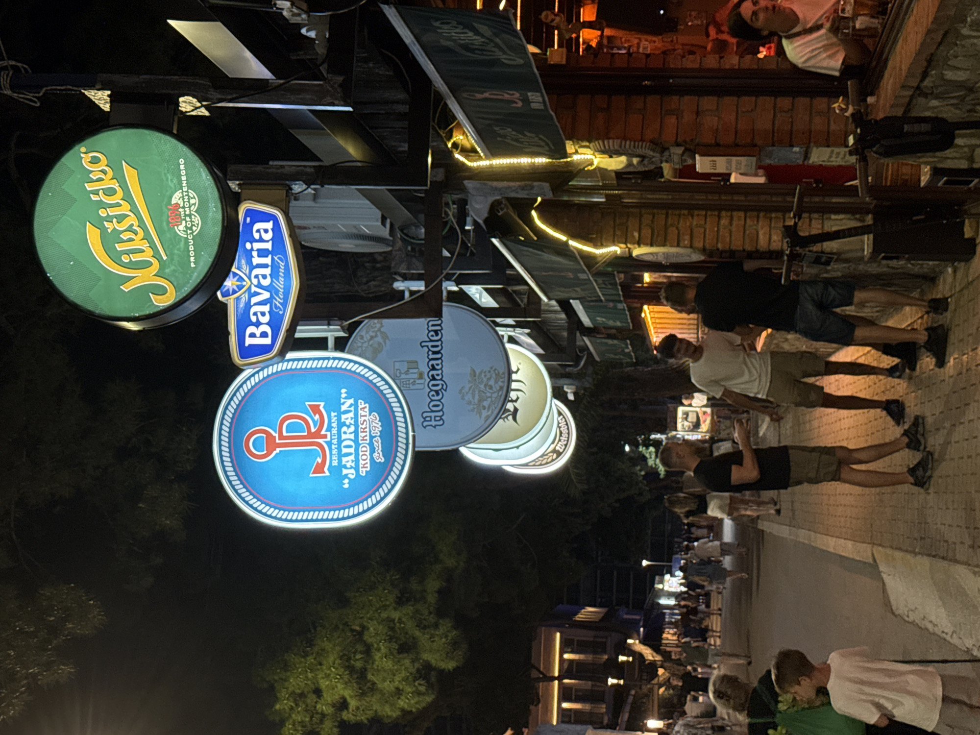 An eye-level, medium shot captures a lively street scene at night in Budva, Montenegro. The foreground is dominated by the sidewalk and a brick-faced building featuring an outdoor seating area. A woman with dark hair, wearing a white t-shirt, is visible through the building's window, holding a glass of beer. Several illuminated signs are prominently displayed above the building, advertising brands such as "Nikšičko," "Bavaria Holland," "Hoegaarden," and "JADRAN KOD KRSTA."
In the middle ground, a paved street is lined with trees and pedestrians. Several people are walking along the street, including a young boy in a white t-shirt and two men in casual attire standing on the sidewalk, engaged in conversation. They are standing near the entrance of the building, with scooters parked nearby. The background reveals more buildings and trees, all illuminated by streetlights and the glowing signs, creating a vibrant and energetic atmosphere. The overall lighting suggests it is nighttime, with warm yellow lights highlighting the outdoor spaces.