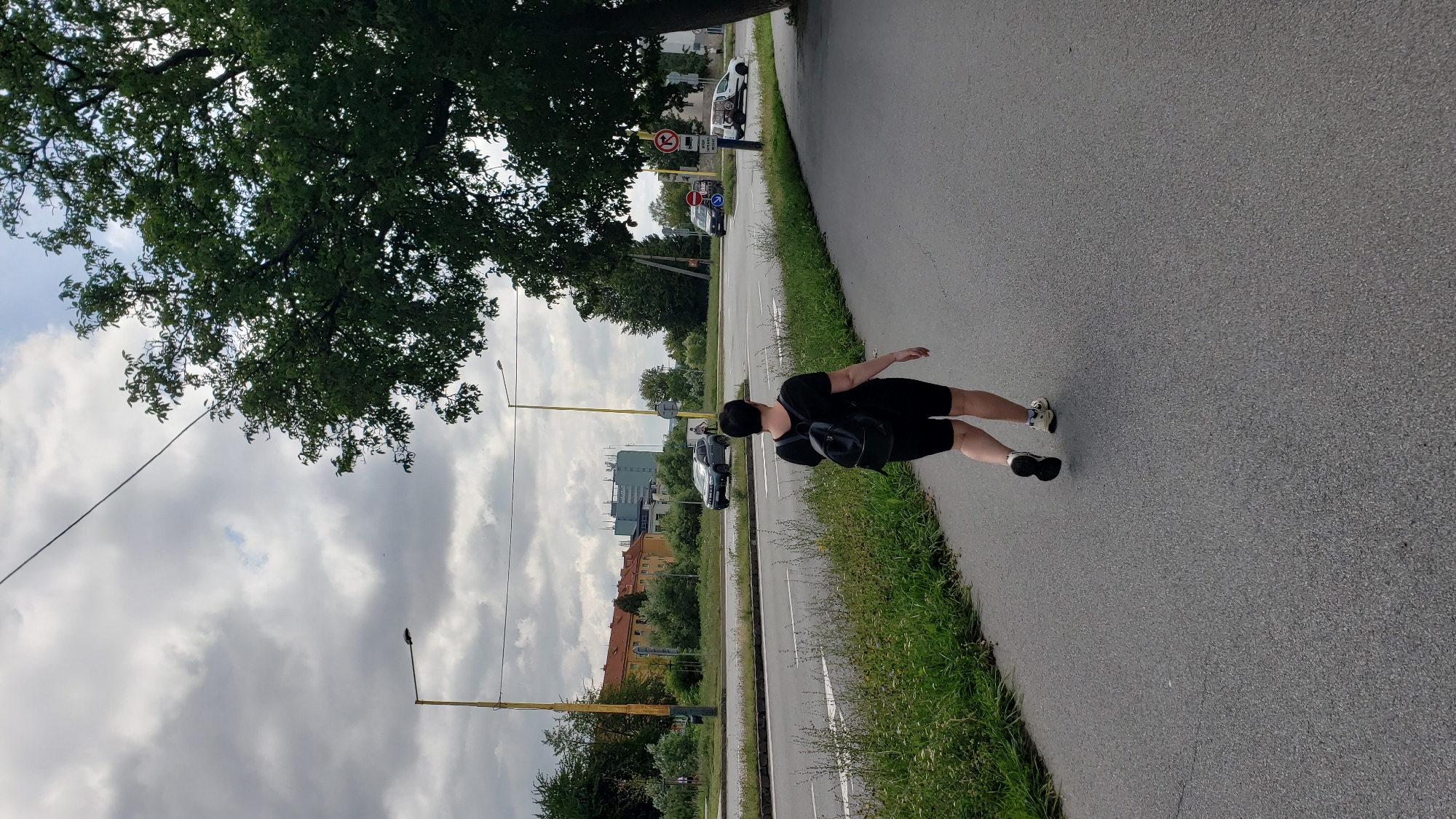 A person is walking away from the camera on a paved sidewalk next to a road with multiple lanes. The person is wearing a black t-shirt, black shorts, a black backpack, and athletic shoes. The sidewalk is bordered by a grassy area with weeds. Across the road, there are more grassy areas, trees, streetlights, and buildings. Some of the buildings appear to be residential, with orange roofs, while others are modern, multi-story structures. Cars are visible on the road, including a dark colored SUV, a dark van, and a white van. Traffic signs are present near the road. The sky is overcast with clouds. This scene is located on the street named "Masarykova" in the city of Prešov, Slovakia.