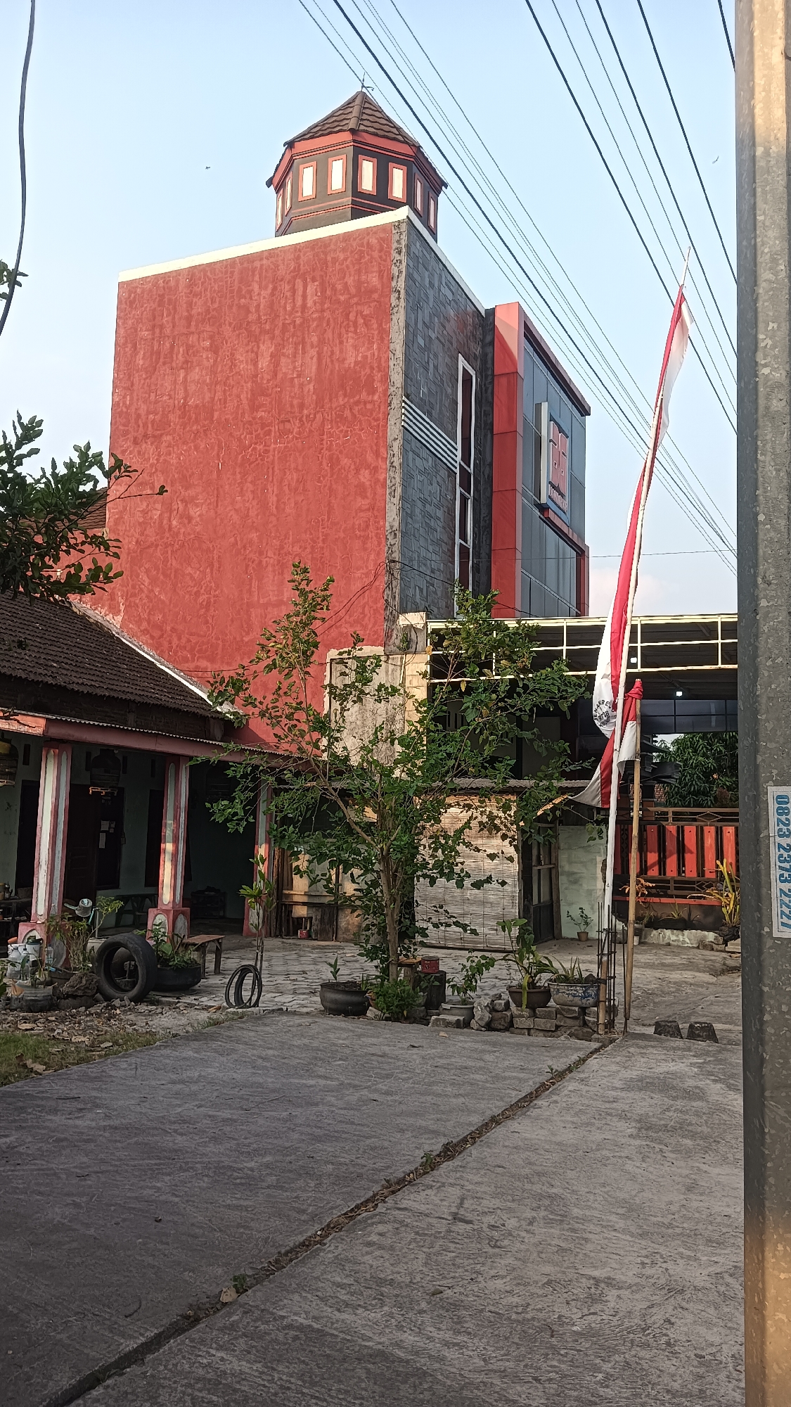 This is an eye-level, outdoor, medium shot of a building in Kedungupit, Indonesia. The building is modern and features red and grey colored walls, with a unique octagonal structure at the top. A red and white Indonesian flag is prominently displayed on the right side of the image, attached to a pole. The foreground shows a concrete pathway leading to the building, with various potted plants and some discarded tires scattered around. A smaller, older building with a tiled roof is visible to the left of the main structure. The sky is clear and light blue, with some power lines visible across the top of the frame. The overall scene suggests a residential or commercial area in a tropical climate.