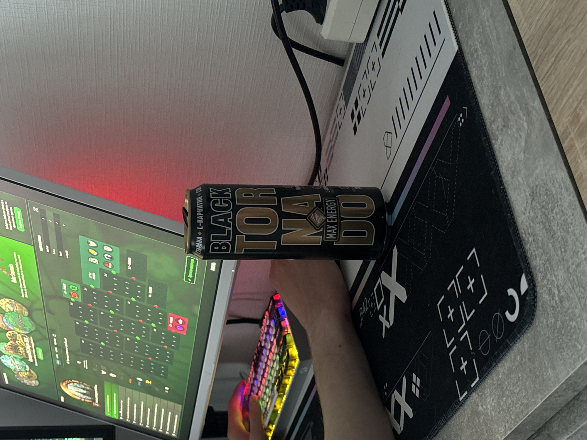 The image shows a person sitting at a desk with a computer monitor displaying a strategy game. A black and gold energy drink can, labeled "Tornado Black Max Energy", is positioned near the monitor. The person's hand is visible near a colorful, illuminated keyboard. A black and white mousepad is under the keyboard. The environment includes a white wall and a gray desk surface. There is no indication of the location being Vsevolozhsk, Russia in the image.