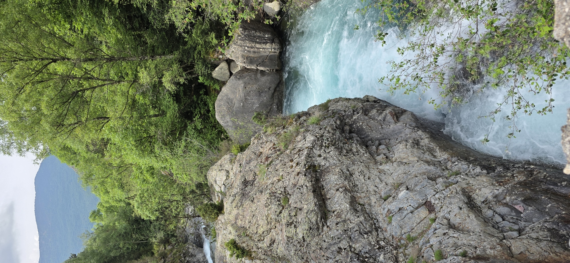 A dynamic natural landscape in Sahún, Spain, captured from an elevated perspective looking down into a river gorge. The central feature is a rapidly flowing river, characterized by its striking turquoise-blue water, which appears foamy white in turbulent areas. Large, rugged grey and brown rock formations and boulders line the riverbanks and form a prominent cliff face in the foreground.
Lush, dense green deciduous trees and various smaller shrubs cover the right bank of the river and extend across the distant mountain slopes. A large, verdant mountain rises in the background on the left side. The scene depicts the natural flow of the river through its rocky environment.
The time of day appears to be daytime, with bright, diffused lighting suggesting an overcast sky or soft sunlight. There are no human subjects or visible text. The powerful current, vibrant water color, and mountainous, forested setting are notable features of this natural environment in the Spanish Pyrenees.