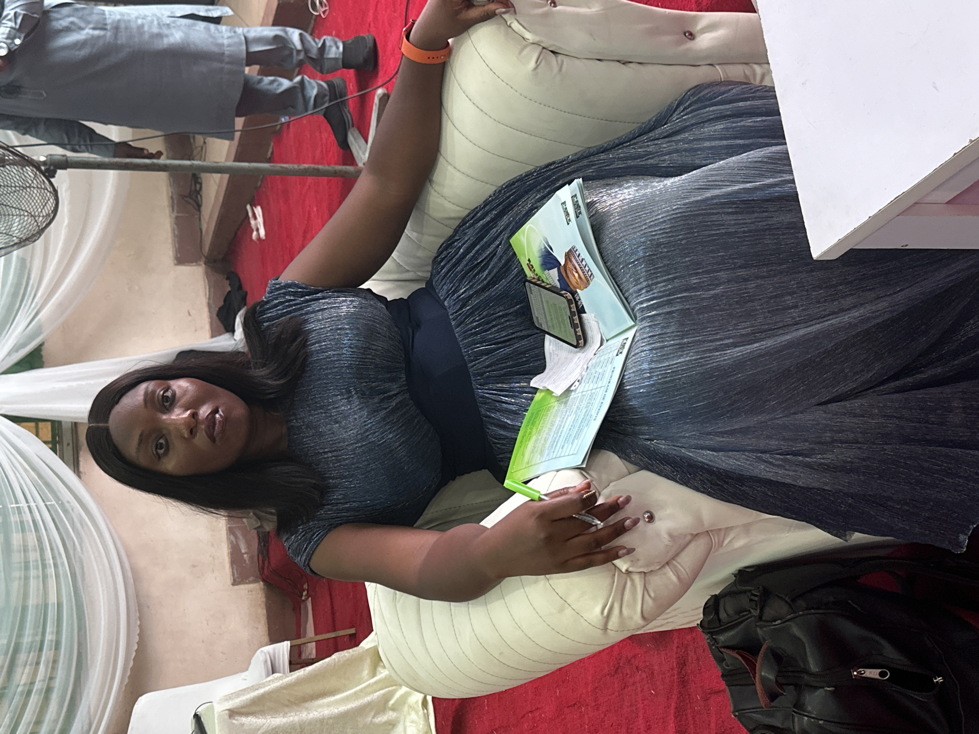 A woman is reclined on a cream-colored cushion, dressed in a sparkling, dark-colored, flowing gown. She has a notebook and a smartphone resting on her dress. To the right of her, a black bag is visible. A white table is partially visible at the top right corner. A fan stands to the left. There is a glimpse of a person wearing a gray outfit at the top left corner. The location is indicated as Unknown, Unknown.