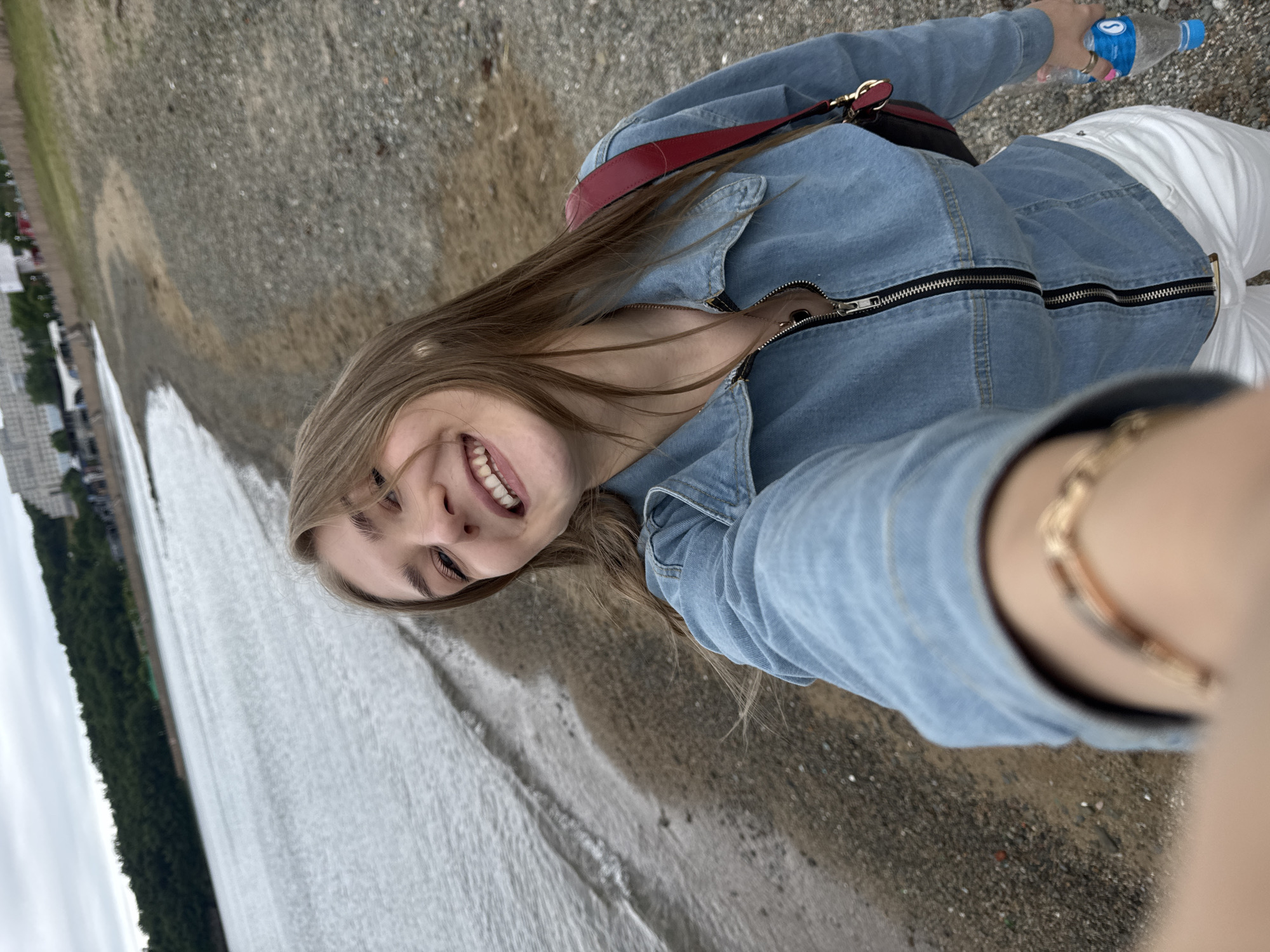 The image shows a young woman taking a selfie on the pebble beach of Russky Island, Russia. She is wearing a blue denim jacket, white pants, and a red purse over her shoulder. The water of the bay is visible to her left, and a forested hill with buildings in the distance is to the left of the bay. She is holding a bottle of water in her right hand.