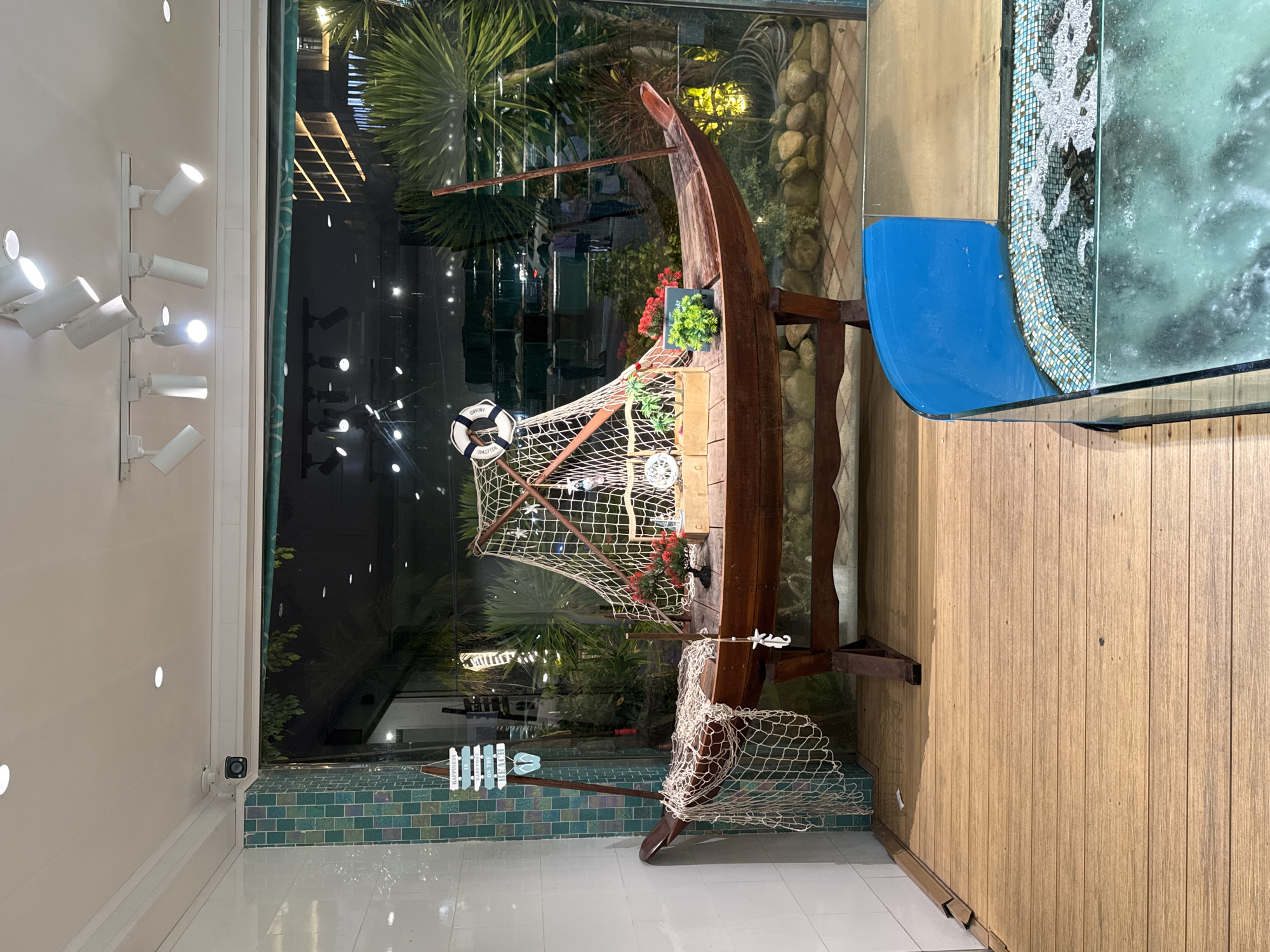 This image depicts a decorative display on a wooden deck in what appears to be a restaurant or shop in Da Nang, Vietnam. The central element is a wooden boat, resembling a sampan, placed on a stand. The boat is adorned with fishing nets, a life preserver with the word "WELCOME" visible, small decorative palm trees, a red floral arrangement, and a white seahorse ornament. To the left of the boat, a wooden sign with the words "HAPPINESS IS BAREFOOT ON THE BEACH" and two flip-flops is attached to a pole. The display is situated in front of a large glass window that looks out onto an outdoor area with lush greenery, palm trees, and some indistinct figures of people walking in the background. The overall impression is a nautical or beach-themed decoration, likely intended to evoke a coastal ambiance. The foreground features a portion of a glass-enclosed aquarium with blue trim and a tiled mosaic border, suggesting the establishment might be a seafood restaurant. The floor is made of weathered wooden planks. The lighting appears to be a mix of interior spotlights and ambient light from the outside.