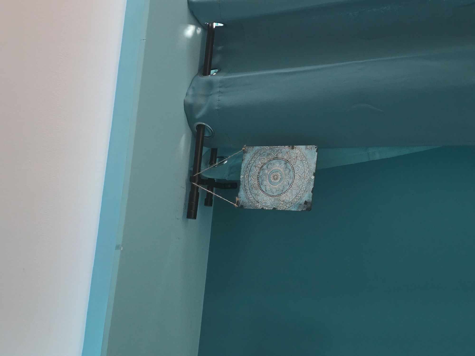 In an interior scene in Malko Tarnovo, Bulgaria, a decorative square plaque with a mandala pattern is suspended by twine from a dark metal curtain rod. The plaque is light blue with intricate white designs. To the right of the plaque, a teal curtain is draped. The ceiling above is painted white, with a band of light blue paint along the upper wall. The overall impression is of a simple, minimalist interior decoration.