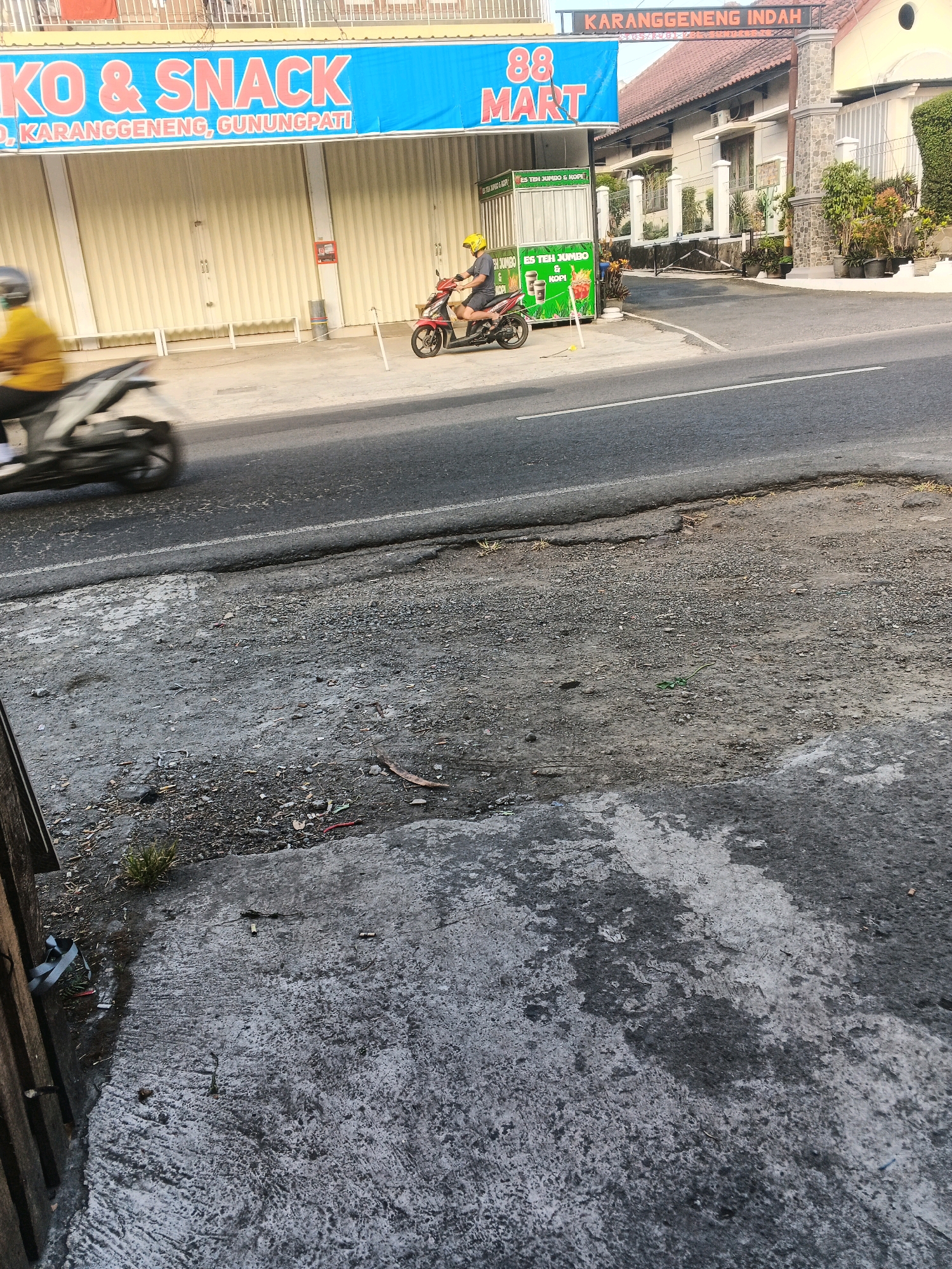 This image captures a street scene in Semarang, Indonesia, specifically along a road in front of a building identified by signage as "88 MART" and "KARANGGENENG INDAH." The building appears to be a storefront with a large, light-yellow rollup door. A blue banner above the rollup door reads "KO & SNACK" and below it, "D, KARANGGENENG, GUNUNGPATI." To the right of the storefront, a smaller green cart displays images of drinks and fries, with text that includes "ES TEH JUMBO & KOPI."
In the foreground, a person wearing a yellow shirt and a yellow helmet is riding a red motorcycle. They are captured in motion, heading away from the viewer. Another person, also on a motorcycle and wearing a yellow shirt and helmet, is in the process of riding past the storefront, also heading away from the viewer, and is blurred due to motion. The street in the foreground is asphalt with visible white lane markings. The side of the road is composed of rough ground and concrete. A wooden fence is partially visible on the far left of the frame. The environment suggests a commercial area in Semarang.
