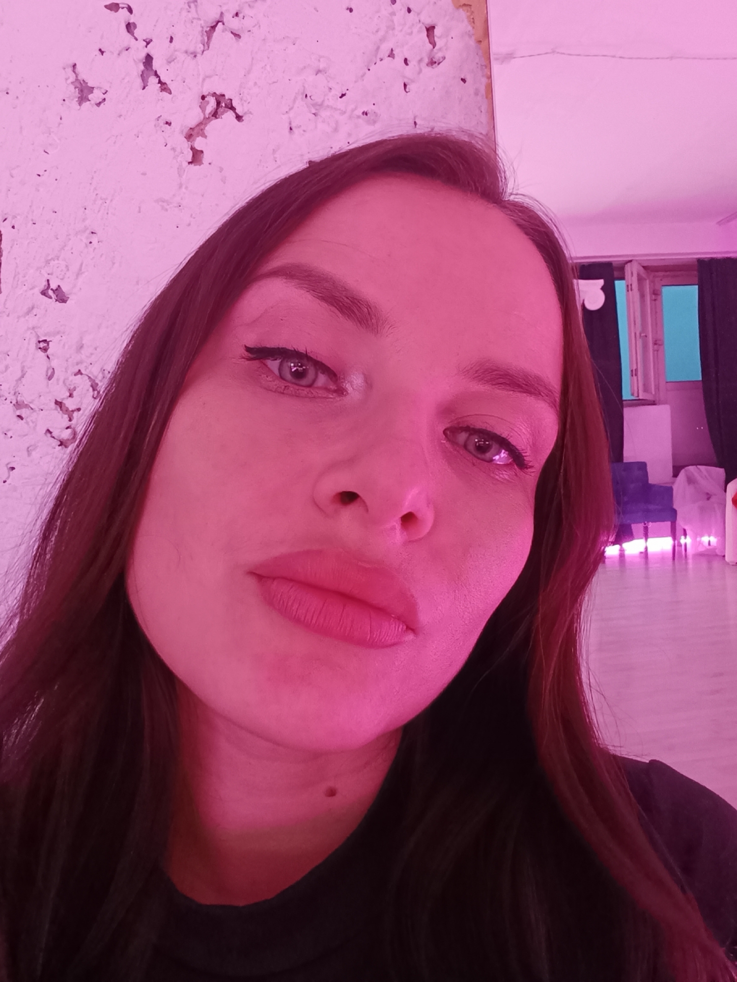 A close-up selfie photograph shows a woman with light skin, dark hair, and light eyes. She is wearing eyeliner and has full lips. A mole is visible on her neck. The lighting is predominantly pink, giving the image a magenta hue. The background includes a white wall with some texture, and a room with a window, dark curtains, a blue chair, and what appears to be neon lighting on the floor. The location is in the city of Unknown, Unknown.