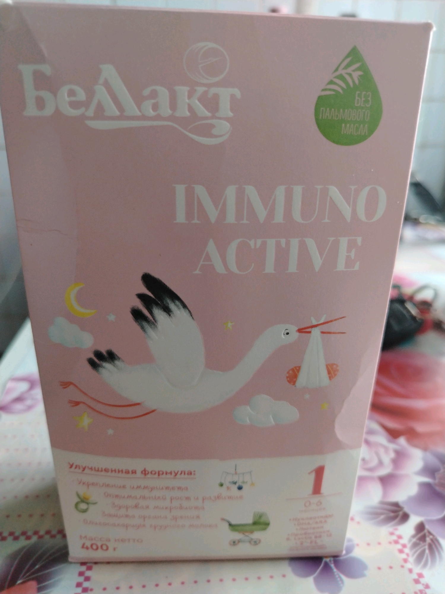 The image shows a pink box of baby formula. The brand name "Белакт" is prominently displayed in white lettering at the top of the box, with a circular logo to its right. Below the brand name, the words "IMMUNO ACTIVE" are written in large white letters.
The box features illustrations of a stork carrying a baby wrapped in a blanket, a crescent moon, clouds, and stars. There is also a green droplet shape on the upper right side of the box containing the text "БЕЗ ПАЛЬМОВОГО МАСЛА" which translates to "WITHOUT PALM OIL."
Towards the bottom left of the box, there is text in Russian that describes the formula's benefits, including "Улучшенная формула" (Improved formula), "Укрепление иммунитета" (Immune system strengthening), "Оптимальный рост и развитие" (Optimal growth and development), "Здоровая микрофлора" (Healthy microflora), and "Защита органов зрения" (Eye protection). Below this, it states "Масса нетто 400 г" (Net weight 400g).
On the right side of the box, a large red number "1" is visible, indicating it is stage 1 formula, suitable for ages 0-6 months.
The box is placed on a surface with a patterned tablecloth featuring flowers in shades of pink, purple, and white. The background is blurred but appears to be an indoor setting, possibly a home. There is no discernible connection to the city of Чавусы, Belarus, as no geographical landmarks or context are provided within the image itself.