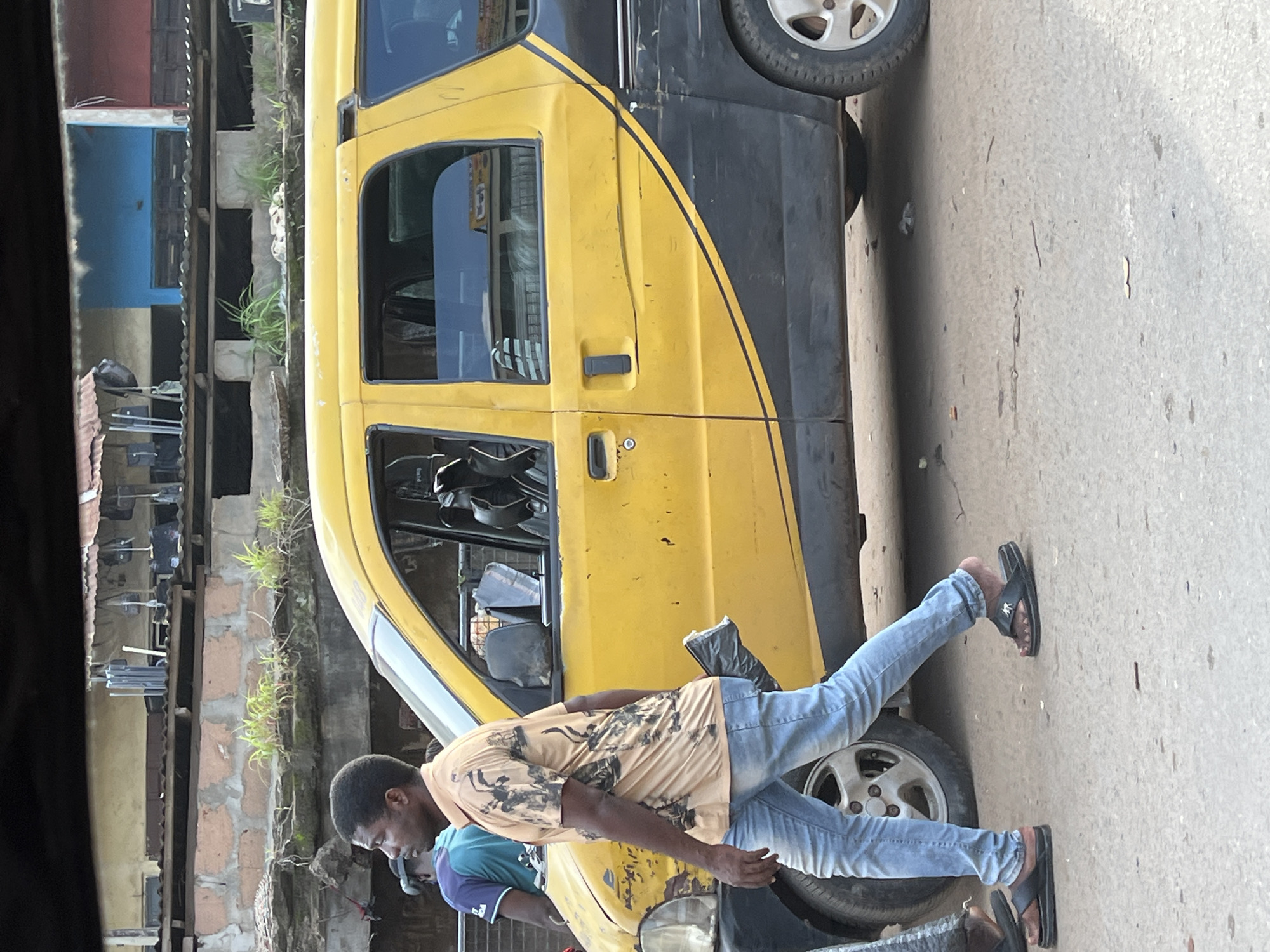 In Nkpor, Nigeria, a yellow and black van is parked on a paved surface. A Black man wearing a tan patterned short-sleeved shirt, blue jeans, and black flip-flops is walking in front of the van. He is holding a black plastic bag in his right hand. Behind him, another person wearing a blue shirt is visible. The background shows buildings with corrugated metal roofs and brickwork.
