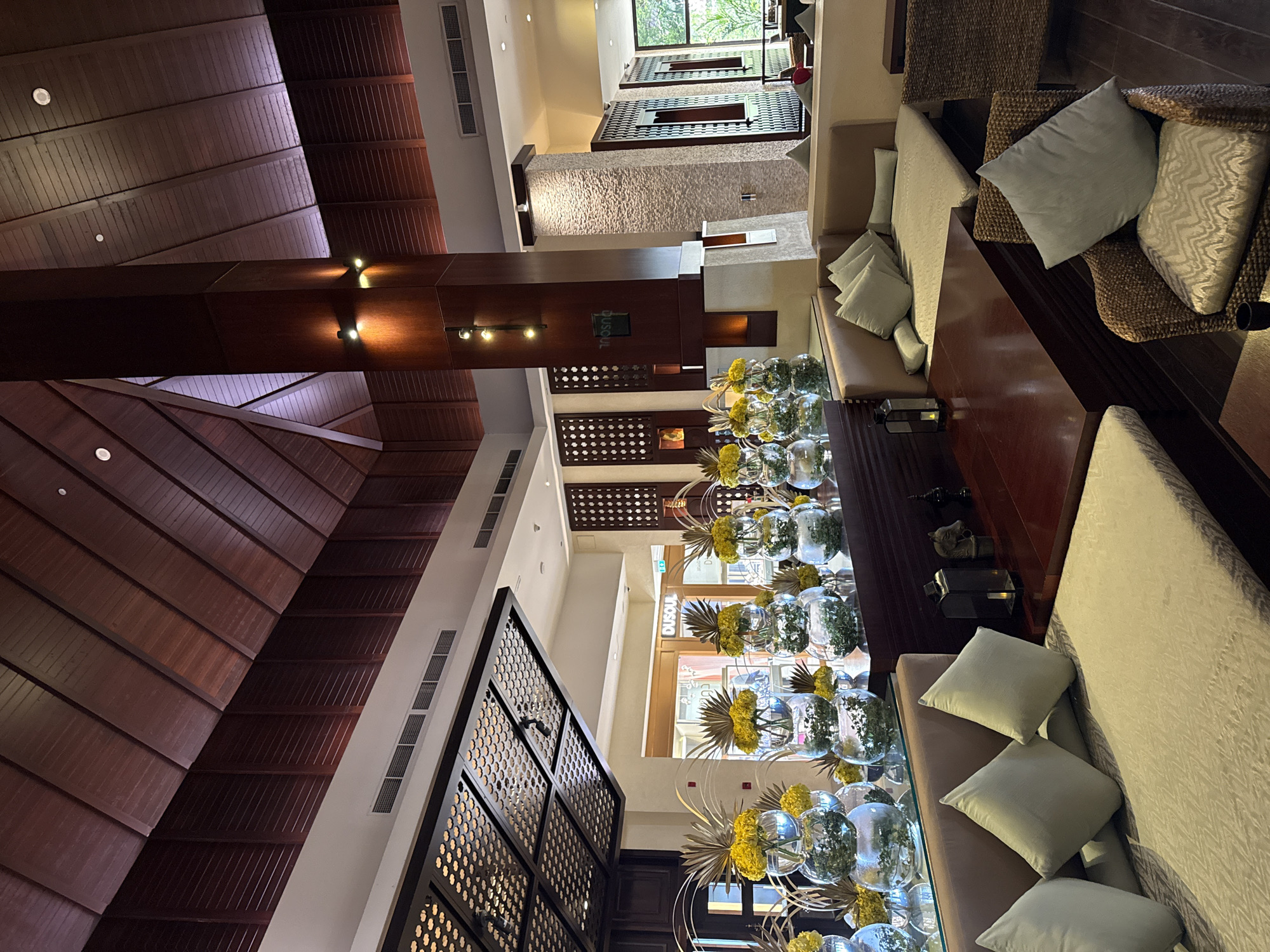 The image depicts the interior of a luxurious hotel lobby in Dubai, United Arab Emirates. The space is characterized by a high, vaulted wooden ceiling with intricate latticework and recessed lighting. A prominent, dark wood pillar stands in the center of the frame, adorned with modern wall sconces that cast a warm glow.
The foreground features a seating area with plush, light-colored sofas and armchairs upholstered in textured fabric. Scattered across the seating are several light grey throw pillows. A dark, polished wooden coffee table sits in front of the sofa, with a minimalist lantern and decorative objects placed on it.
On a reflective surface behind the seating area, a series of elaborate floral arrangements are displayed. These arrangements consist of stacked clear glass vases filled with greenery and topped with vibrant yellow flowers and golden, fan-shaped foliage.
The background reveals the hotel's storefronts and entrance, with one prominently displaying the signage "DUSOUL". The walls are decorated with patterned wooden panels and textured stone accents. Large windows offer glimpses of the outdoor environment, suggesting a tropical or garden setting. The overall ambiance is one of sophisticated elegance and comfort, typical of high-end hospitality establishments in Dubai. There are no people visible in the image.