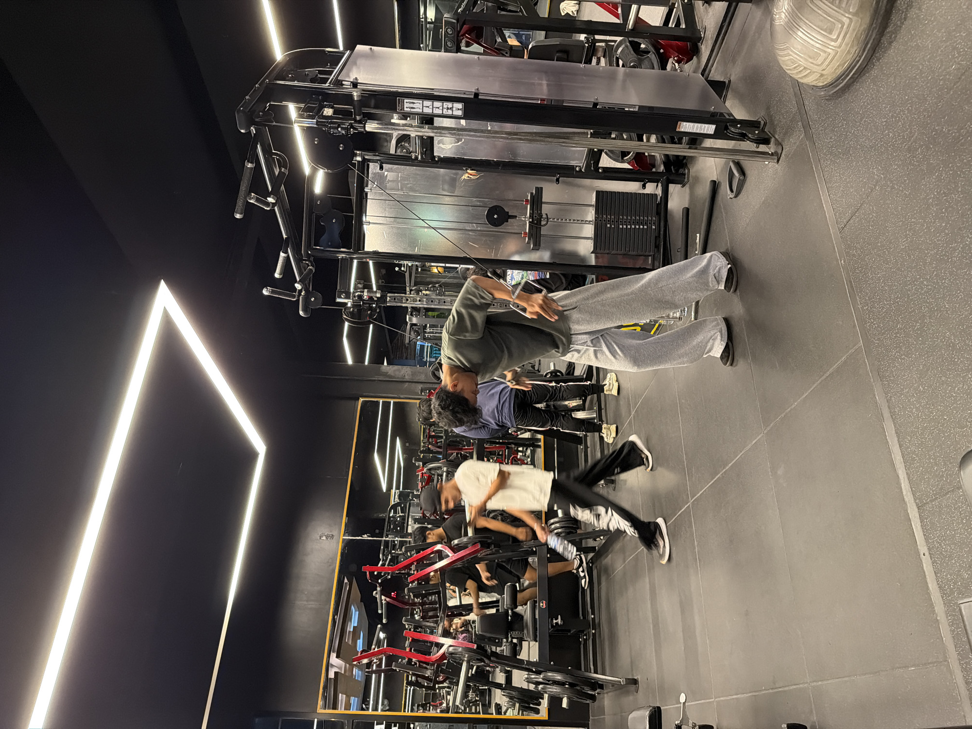 The image shows the interior of a gym in Denpasar, Indonesia. Several people are engaged in various exercises using weight machines and cable stations. The gym features a large mirror reflecting the exercise area, exposing several people exercising. The gym has black walls accented by geometric LED lighting fixtures. The floor is covered with gray tiles. Weight machines with red and black frames are visible in the reflection. In the foreground, a person wearing gray sweatpants and an olive-green t-shirt is using a cable machine.