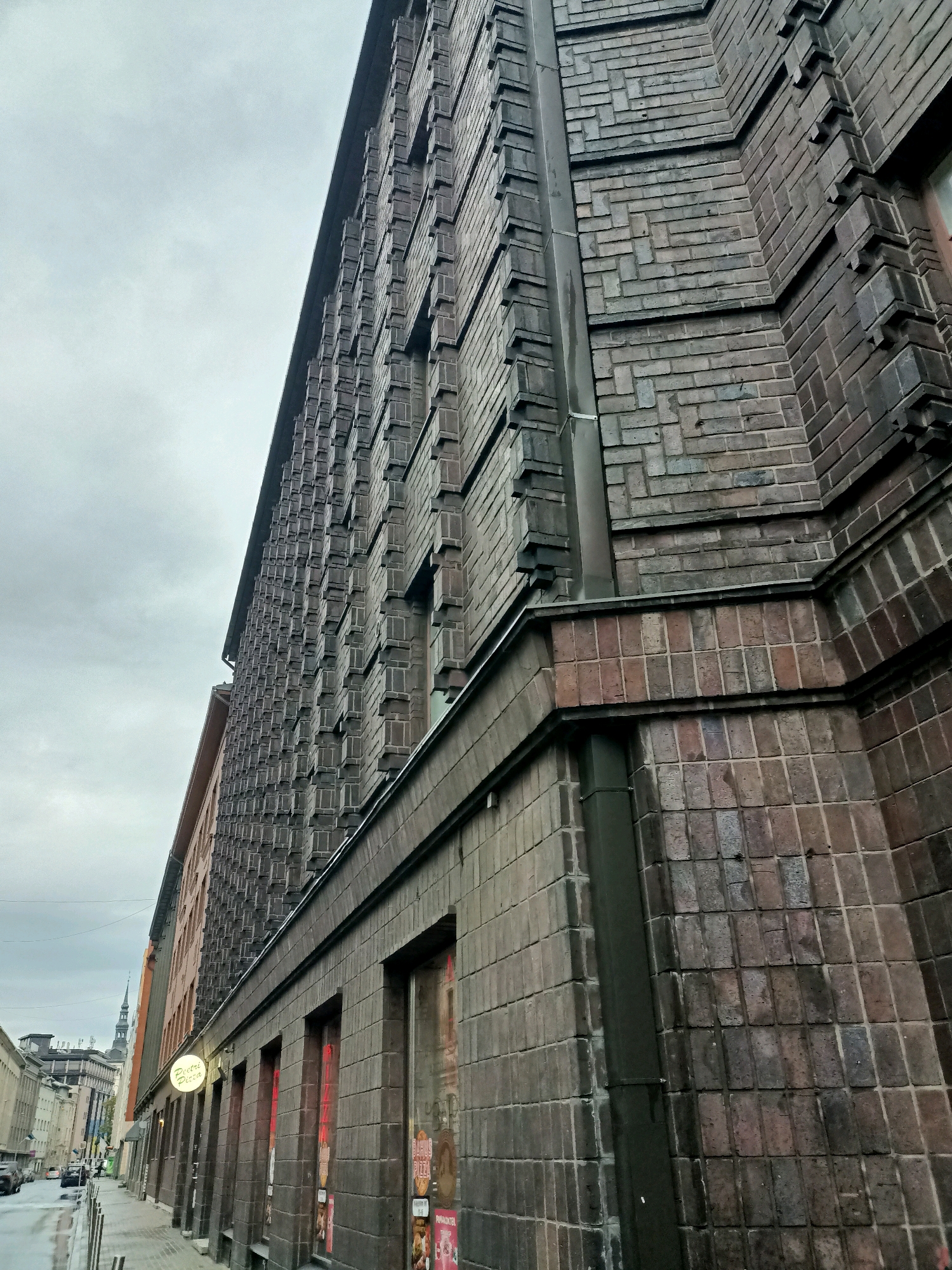 The image captures a view of a building facade on a street in Tallinn, Estonia. The building is constructed with dark, rectangular bricks and features a distinctive, stepped architectural design along its upper sections. The facade is largely unadorned, with a series of windows visible at street level, some displaying advertisements. A drainpipe runs vertically along the side of the building. The street in front of the building is paved with cobblestones and appears wet, suggesting recent rain. A sidewalk lines the street, with a few streetlights visible. In the distance, other buildings and a church spire can be seen, characteristic of an urban environment. The sky is overcast, contributing to the muted tones of the scene. There are no people or vehicles clearly depicted in the foreground, though the presence of a street implies traffic. The overall impression is one of an older, European city on a cloudy day.