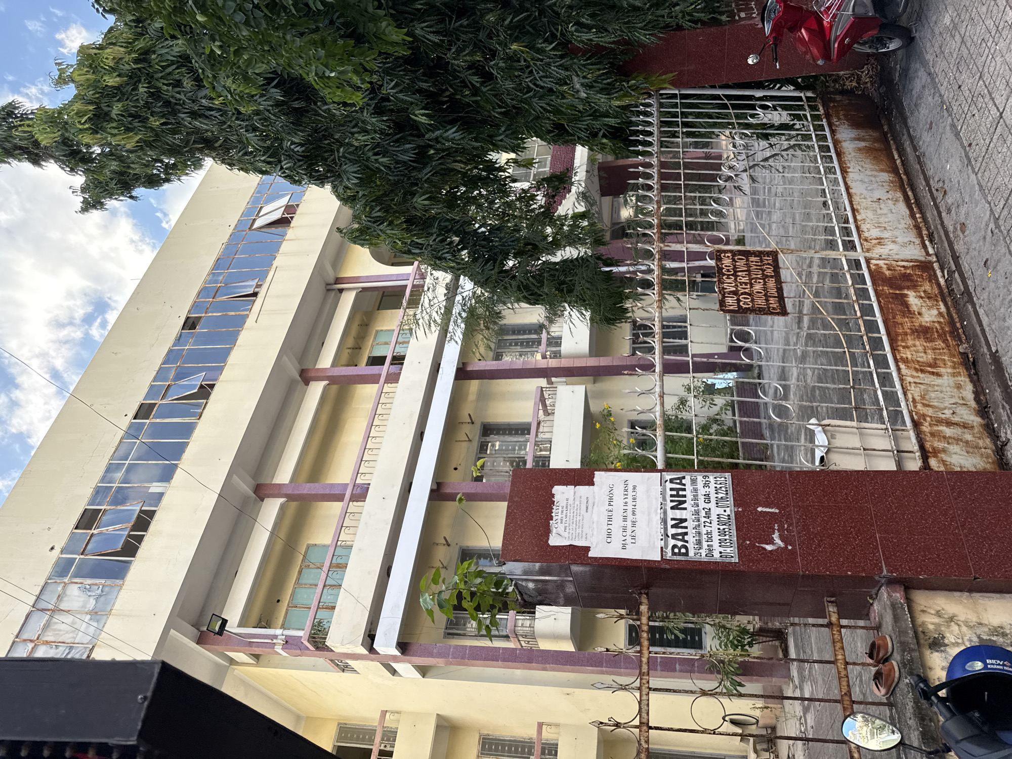 A multi-story building with yellow walls and a light-colored facade stands under a cloudy sky in Nha Trang, Vietnam. The building has multiple windows, some of which are open and appear to be covered with rust-stained metal frames. Several balconies with pink railings are visible, and a large tree with dark green leaves partially obscures the building. A metal fence, also showing signs of rust, runs along the front of the property. Two pairs of children's shoes are placed on a ledge near the fence. A sign attached to a red pillar by the fence reads "BAN NHÀ" (House for Sale) with Vietnamese text and a phone number. Another sign further down the fence, in Vietnamese, warns against parking. A red motorcycle is parked to the right of the fence. The ground in front of the fence is paved with gray tiles.