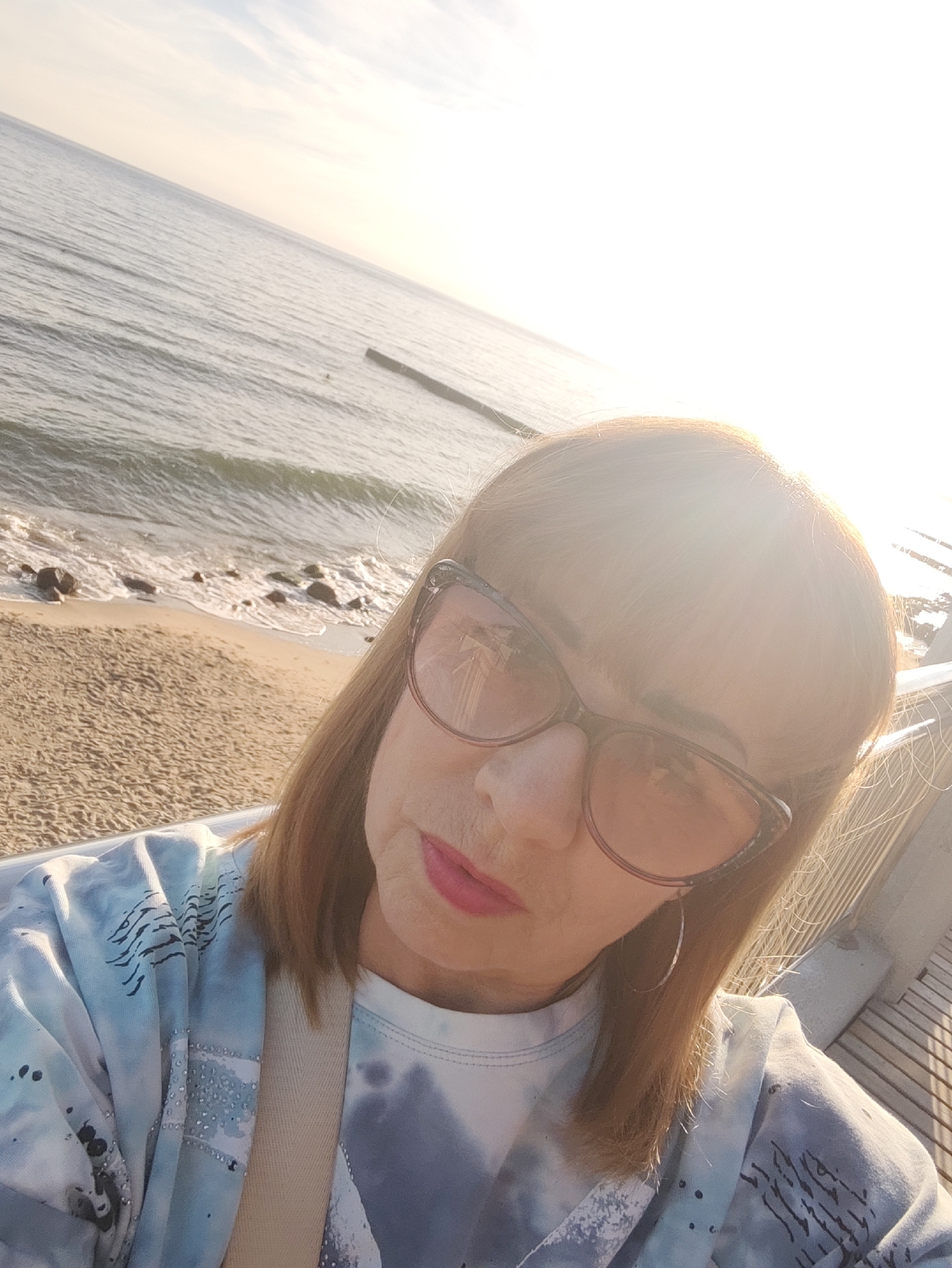 A selfie taken outdoors shows a middle-aged woman with auburn hair and bangs, wearing large, brown-tinted cat-eye glasses and hoop earrings. She has on a tie-dye sweatshirt with a beige strap visible across her shoulder. Her lips are painted pink. She is on a sandy beach, with waves gently breaking on the shore. The water appears calm and reflects the bright, slightly hazy sky. In the distance, a wooden structure or breakwater is visible in the sea, and a pier or boardwalk runs alongside the woman. The sun is low in the sky, casting a warm glow and creating lens flare. The scene is likely Svetlogorsk, Russia, given the characteristic wooden piers and the general aesthetic of the Baltic Sea coast.
