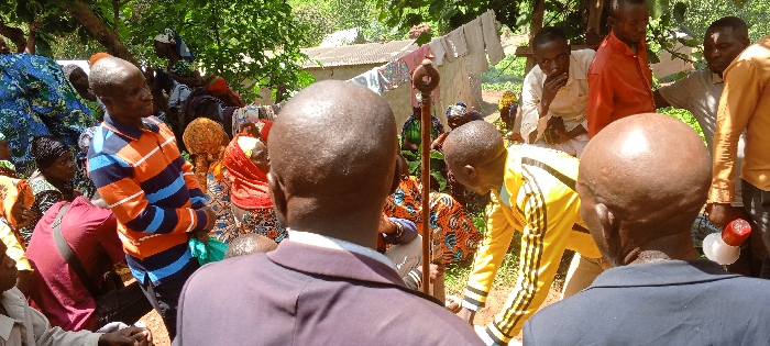 A group of people gathers outdoors. Some sit or kneel, wearing colorful, patterned clothing, while others stand. At least two men in suits are visible from the back. A man in a yellow tracksuit bends forward. In the background, a clothesline with hanging clothes is seen, and a building with a corrugated metal roof peeks through the trees. A tall wooden staff with a circular ornament stands out amidst the crowd.