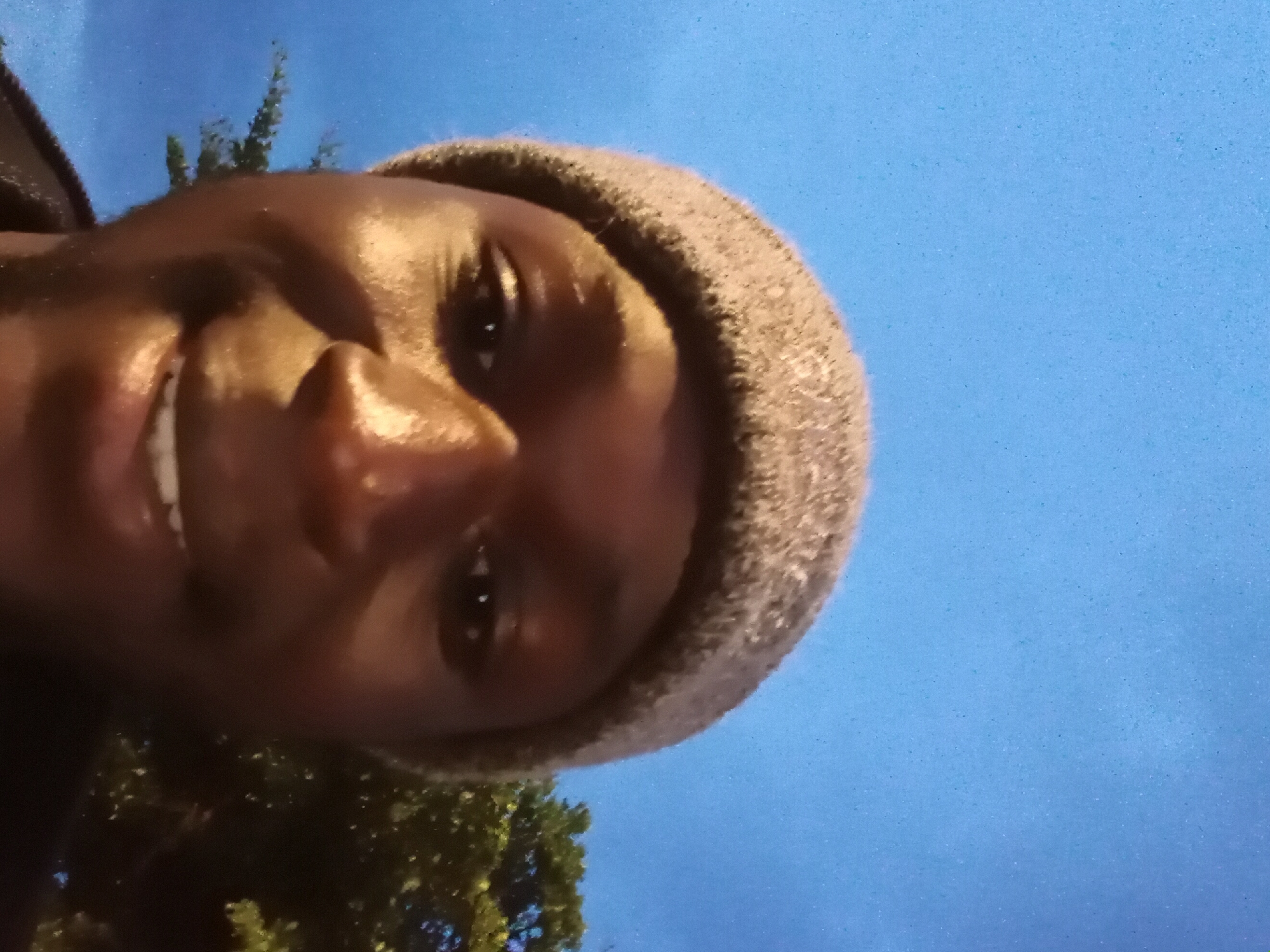 The image is a close-up selfie of a dark-skinned person wearing a grey beanie. The person's face is the primary focus, with their eyes looking slightly upwards. Their mouth is slightly open, revealing teeth, and they appear to be smiling. The background consists of a clear blue sky and some dark green foliage on the right side, suggesting an outdoor setting. The lighting suggests it may be dusk or dawn. The image is taken at an upward angle, characteristic of a selfie taken from a lower perspective.