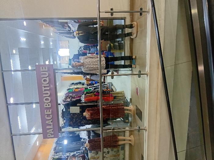 The image shows the storefront of Palace Boutique, located in the city of Pridelands, Kenya. The storefront is visible through a large glass window, showcasing a variety of clothing items displayed on mannequins. Three mannequins are visible, two dressed in women's clothing and one in men's clothing. There is one person standing inside the shop. The person, a young man wearing a light-colored patterned shirt and dark pants, is likely a customer browsing the selection. The glass window has a metal railing in front of it, which is also visible in the foreground. The floor in front of the storefront is tiled, and there is an escalator leading up to the next floor. The escalator is made of metal and has a glass railing.