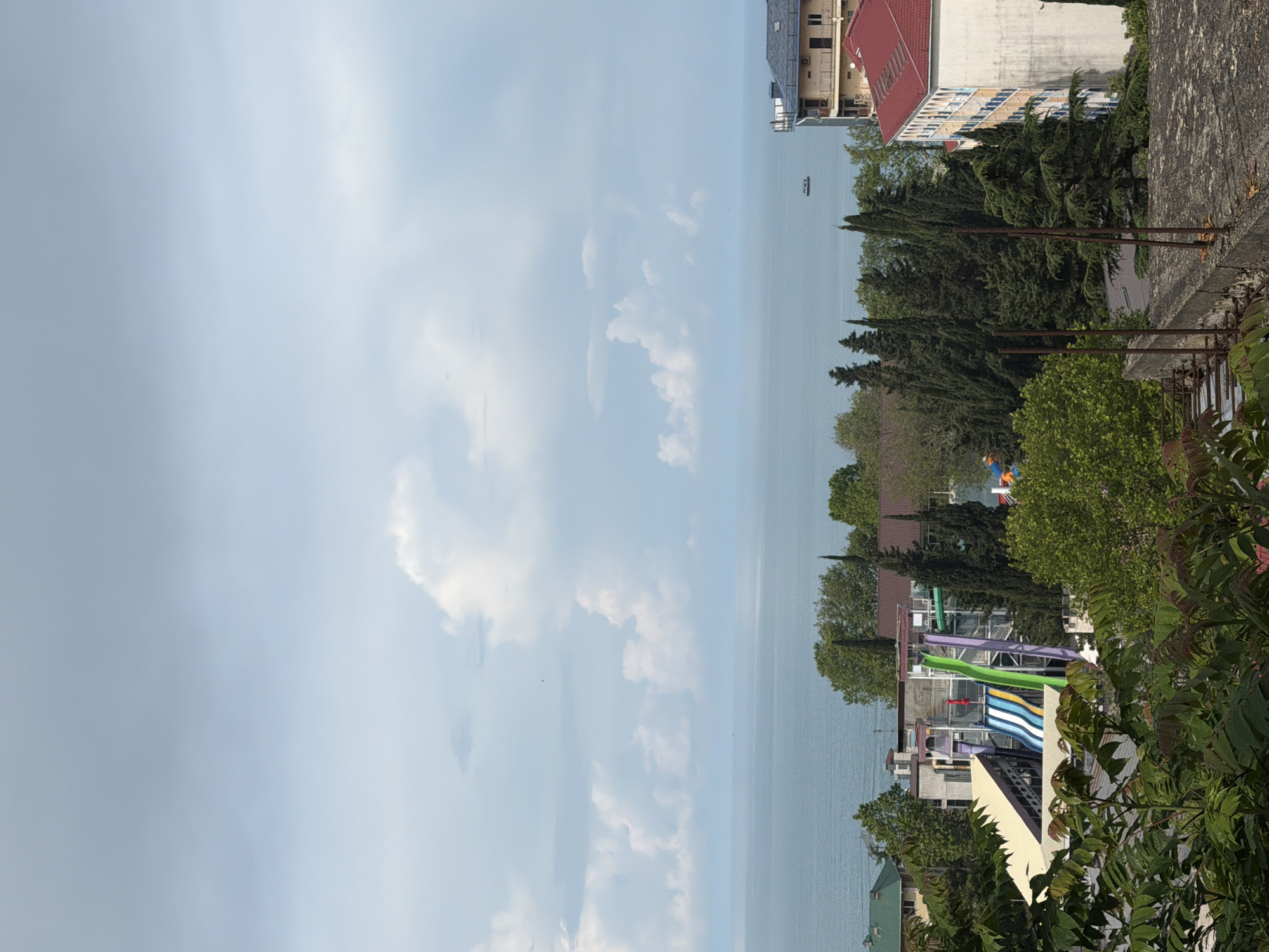 An elevated, wide-angle, eye-level shot captures a coastal city scene with a clear blue sky filled with scattered white clouds. The horizon is marked by the vast, calm expanse of the sea, which transitions into a lighter blue hue near the sky. In the foreground, lush green trees and foliage frame the view, partially obscuring a concrete structure with visible rebar.
Dominating the middle ground is a water park with several colorful slides, including a prominent green slide and blue and white striped slides. Adjacent to the water park are multi-story buildings with beige and white facades, some featuring balconies. One building on the right has a distinctive red roof. Evergreen trees with tall, slender forms are interspersed throughout the landscape.
Further out in the sea, a small, dark boat can be seen. The buildings in the city appear to be hotels or residential complexes, indicative of a resort town. The overall atmosphere is serene, with the natural beauty of the sea and sky contrasting with the urban and recreational elements.
This scene is likely located in Sochi, Russia, a popular resort city on the Black Sea coast known for its beaches, water parks, and surrounding mountainous terrain. The combination of the seaside location, the presence of a water park, and the architectural style of the buildings are characteristic of Sochi's tourist infrastructure.