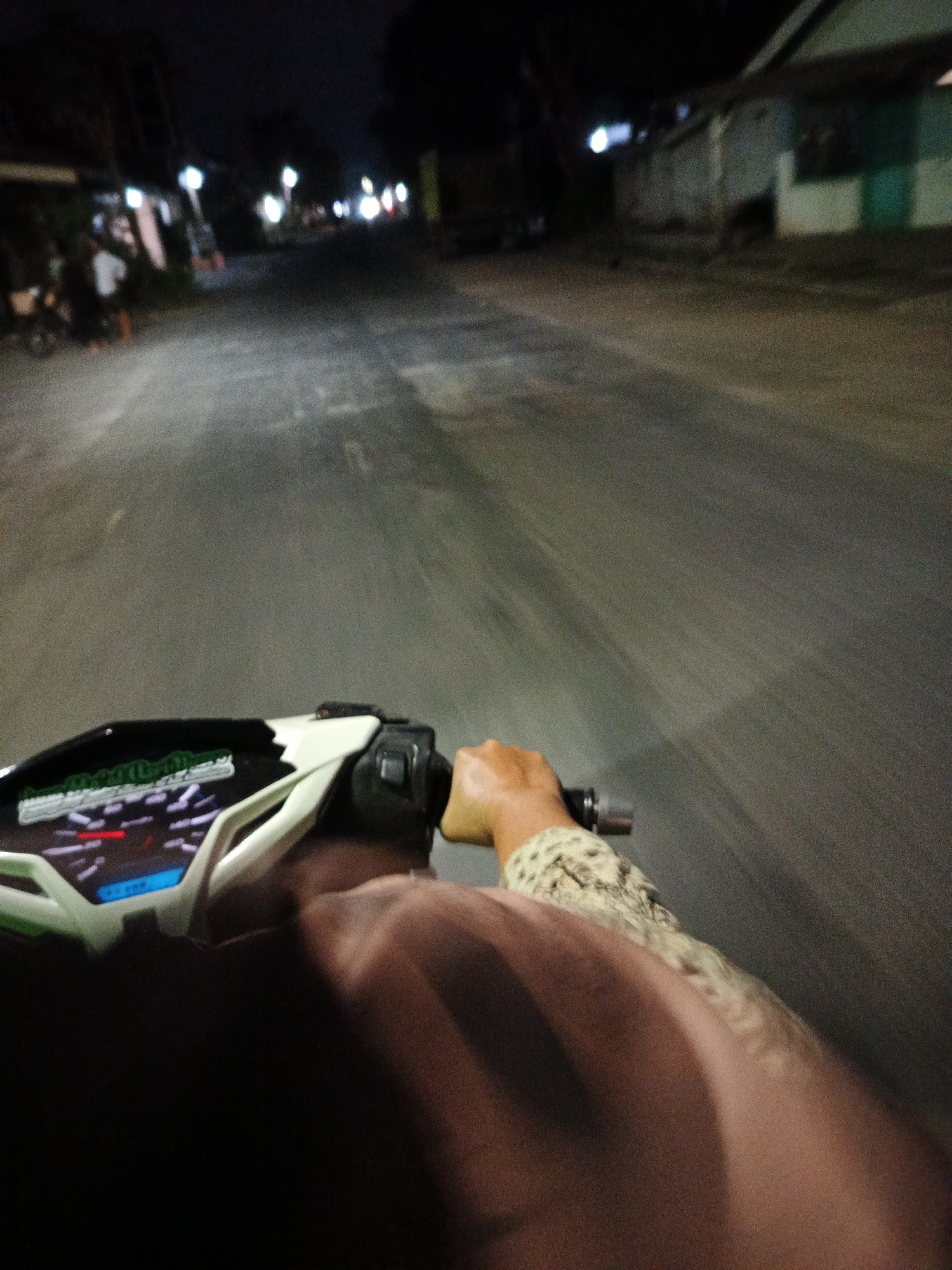 The image captures a first-person perspective of someone riding a motorcycle at night on a paved road. The rider's hands are visible, one gripping the handlebars of the motorcycle, which features a digital dashboard with a red needle indicating speed. The rider is wearing a patterned long-sleeved shirt or jacket. The environment appears to be a street in a city, likely Sumbersari, Indonesia, judging by the presence of buildings and streetlights. The road is visible in the foreground and stretches into the distance, with a slight blur indicating motion. Buildings with lights are visible on the sides of the road, suggesting a populated area. The lighting conditions are dim, consistent with nighttime.