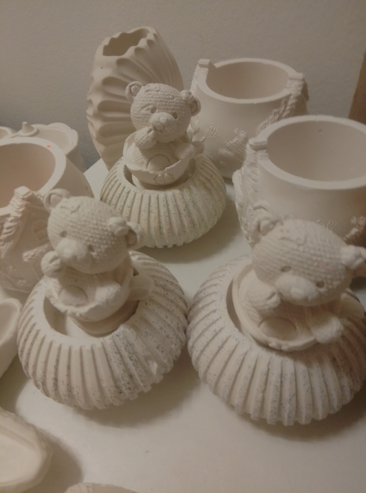 The image shows a collection of unpainted ceramic pieces, primarily featuring teddy bear figurines seated in ribbed bowls or containers. These ceramic items are arranged on a white surface, likely a table or shelf, against a plain, off-white background. The lighting is soft, casting subtle shadows and highlighting the textured surfaces of the unglazed pottery. There are no people or animals present in the scene. The overall impression is one of a crafting or hobbyist's workspace, possibly preparing items for firing or painting. The location of Wedel, Germany, is not discernible from the visual information provided in the image.