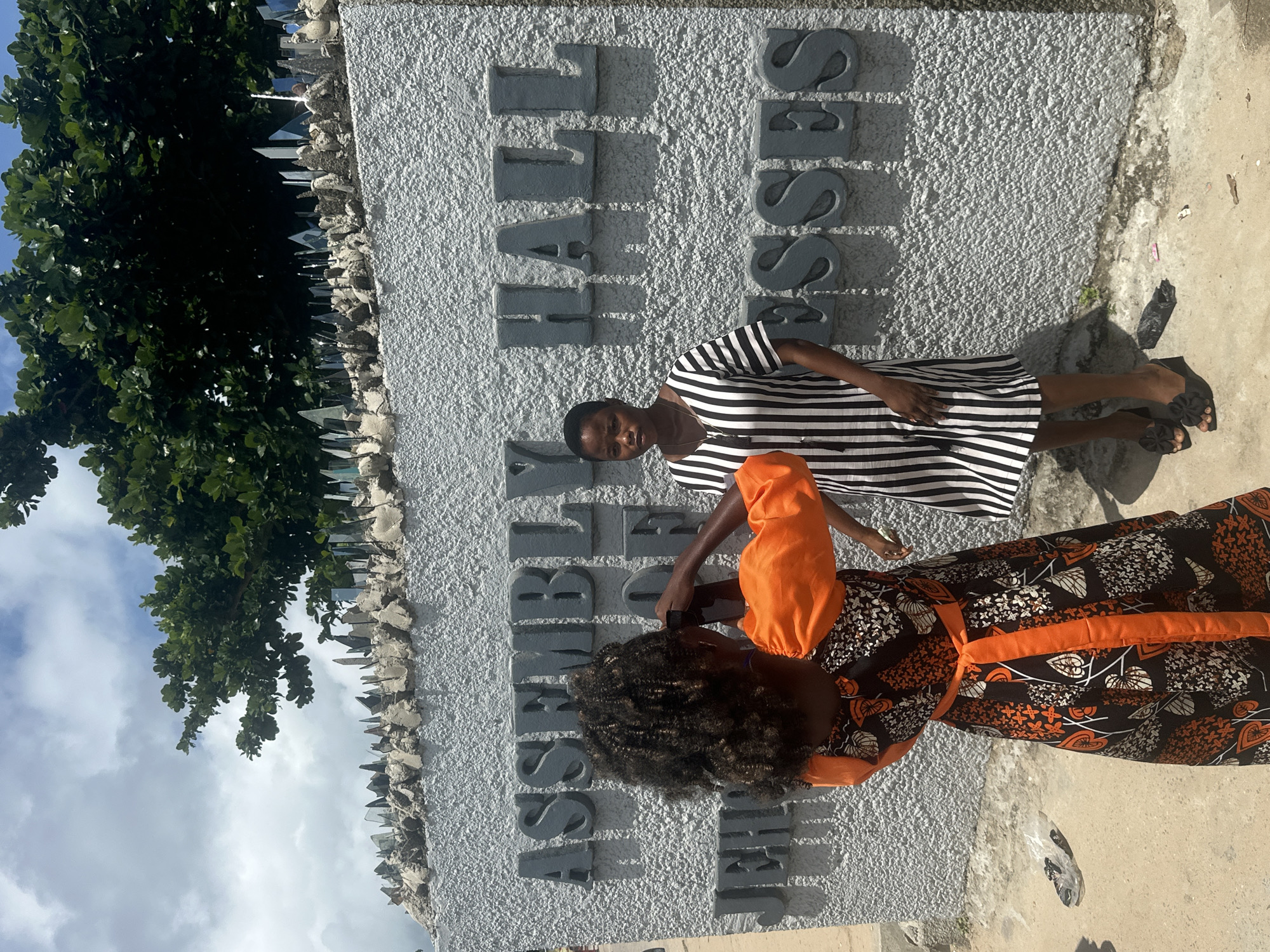 Two young women stand in front of a textured grey concrete wall with large, raised grey letters spelling "ASSEMBLY HALL OF JEHOVAH'S WITNESSES." The woman on the left, wearing a black and orange patterned dress with puffed sleeves and an orange belt, is turned away from the camera and appears to be holding a phone up to take a picture of the woman on the right. The woman on the right stands facing forward, looking towards the camera, and wears a black and white vertically striped dress and black wedge sandals. Behind the wall, lush green foliage is visible, and the top of the wall is lined with broken glass shards embedded in cement, likely for security. The ground in front of the wall is a mixture of concrete and dirt, with some debris scattered about. The sky above is partly cloudy. The location is identified as Itoga, Nigeria.