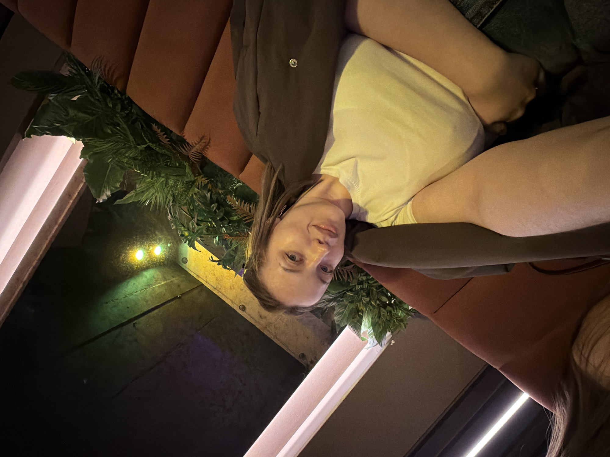 A young adult female is depicted in an indoor setting, likely a restaurant or lounge in Moscow, Russia. The image is rotated 90 degrees counter-clockwise, showing the woman lying or leaning on upholstered seating. She has light brown hair and is wearing a white t-shirt and a dark brown or grey jacket. Her head is slightly tilted, and she looks towards the camera with a neutral to soft expression.
The background features lush artificial green foliage, resembling ferns and other leafy plants, providing a decorative element. She is seated on a large, orange-brown upholstered bench or booth. To her left (top in the rotated image), a dark wall or structure is visible with two small, bright yellowish-green lights, possibly decorative or embedded in the wall. Another rectangular light fixture, likely an LED strip, runs along the bottom edge of the image (right side in the rotated view). The lighting is dim and atmospheric, suggesting nighttime or an evening event. No text is visible in the image.