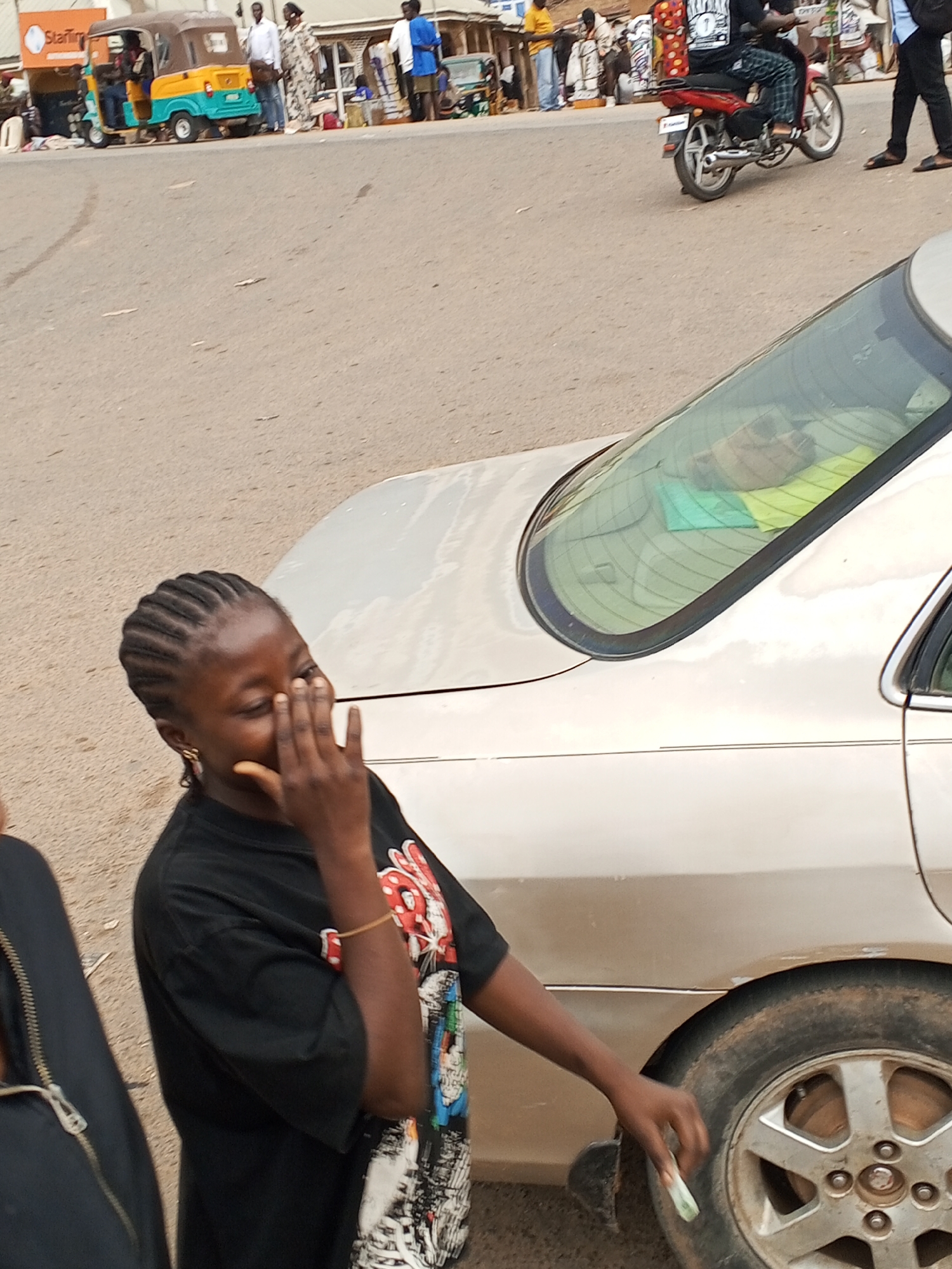 A young woman stands by a gold-colored car near a street in Gwong, Nigeria. The car's rear window is visible with items inside. In the background, there is a three-wheeled vehicle, people standing on the street, and a parked motorcycle. The young woman has her hand to her face. She is wearing a black t-shirt and braided hair.