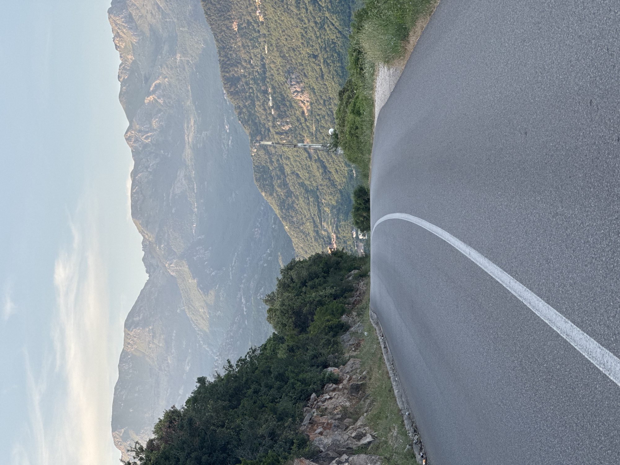 The image shows a paved road with a solid white line marking its edge, winding along a mountain slope in Dubraljevina, Montenegro. The road is bordered by rocky terrain with dense green vegetation on one side. Beyond the vegetation, a mountainous landscape extends into the hazy distance. A tall, thin metal communication tower is visible on the adjacent mountain. The sky is light and overcast.