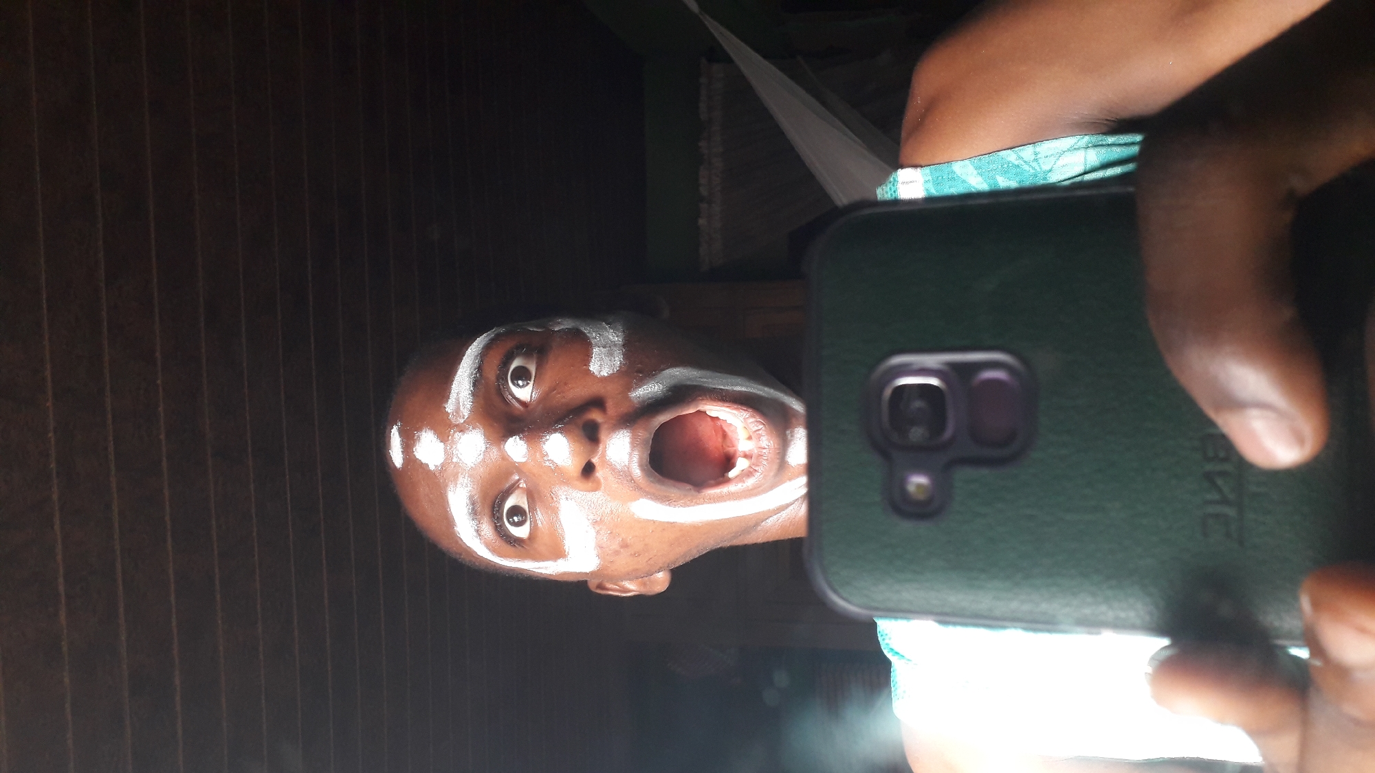 A dark-skinned person with white face paint stands indoors, holding a dark green smartphone to take a selfie. The person's mouth is open in a wide expression. The background includes vertical wooden slats and a white mosquito net. The location is Nkpor, Nigeria.