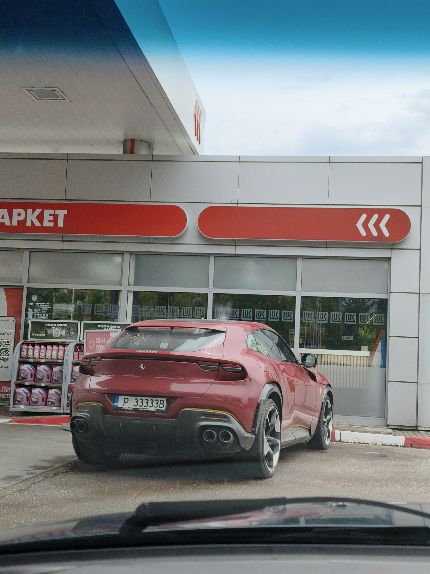 The image captures a maroon Ferrari Purosangue parked at a LUKOIL gas station. The car has yellow accent lines. The license plate reads "P 33333B" with the Bulgarian flag and "www.axlesviat.bg" at the bottom. The gas station has a white exterior, with "APKET" written in red above the entrance and a sign with three left-pointing arrows. Shelves of motor oil are visible to the left of the vehicle. The photograph is taken from inside another vehicle, with part of the dashboard visible at the bottom. The sky is overcast. The city is Unknown, Unknown.