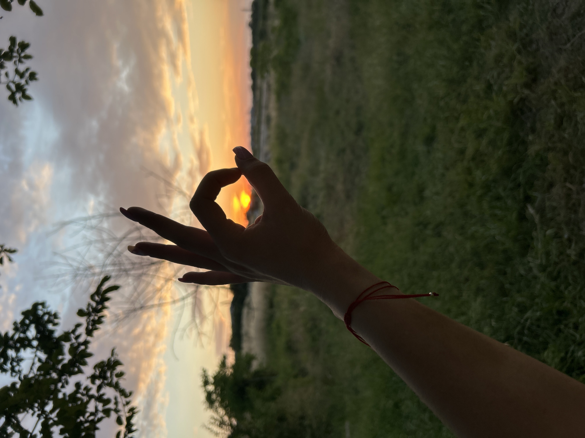 A person's arm, adorned with a red string bracelet, extends into the frame, framing a sunset with their hand in an "OK" gesture. The sun, a bright orange orb, is positioned just above the horizon, casting a warm glow across the sky which is filled with soft, cotton-candy clouds painted in hues of orange, pink, and blue. The background is a blurred expanse of green grass and distant trees, suggesting an outdoor setting, possibly a field or meadow. The scene is captured during the golden hour, emphasizing the soft, diffused light of dusk. The location is not specified.